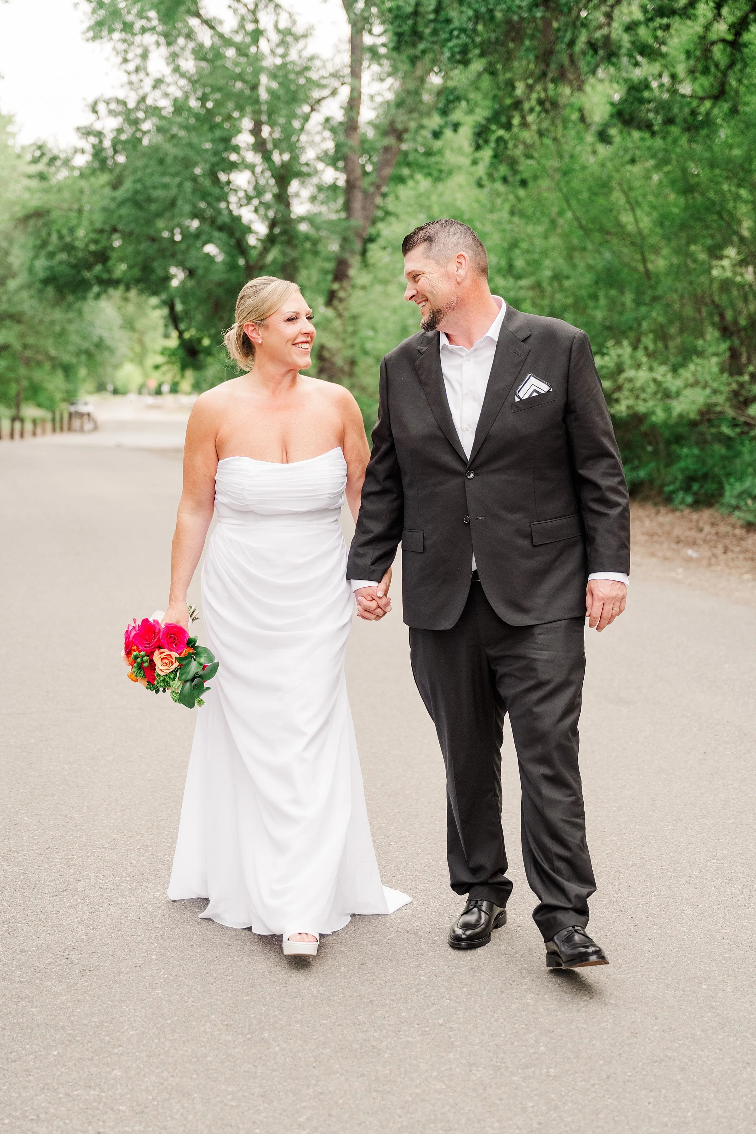 A couple walks hand in hand down a tree-lined path, the woman in a white strapless wedding dress with a bouquet, and the man in a black suit, both smiling warmly.