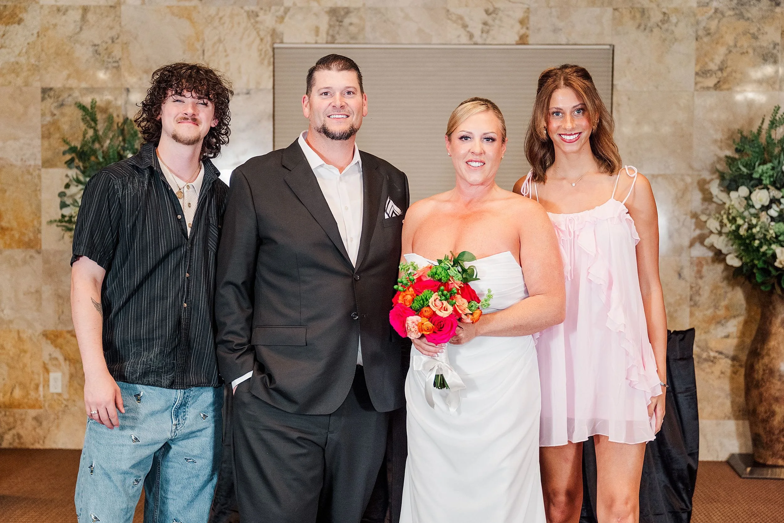 A smiling bride in a white gown holds a vibrant bouquet beside a groom in a black suit. Flanked by a casually dressed man and a woman in a pink dress, the group stands indoors against a marble wall with plants, projecting happiness and celebration.