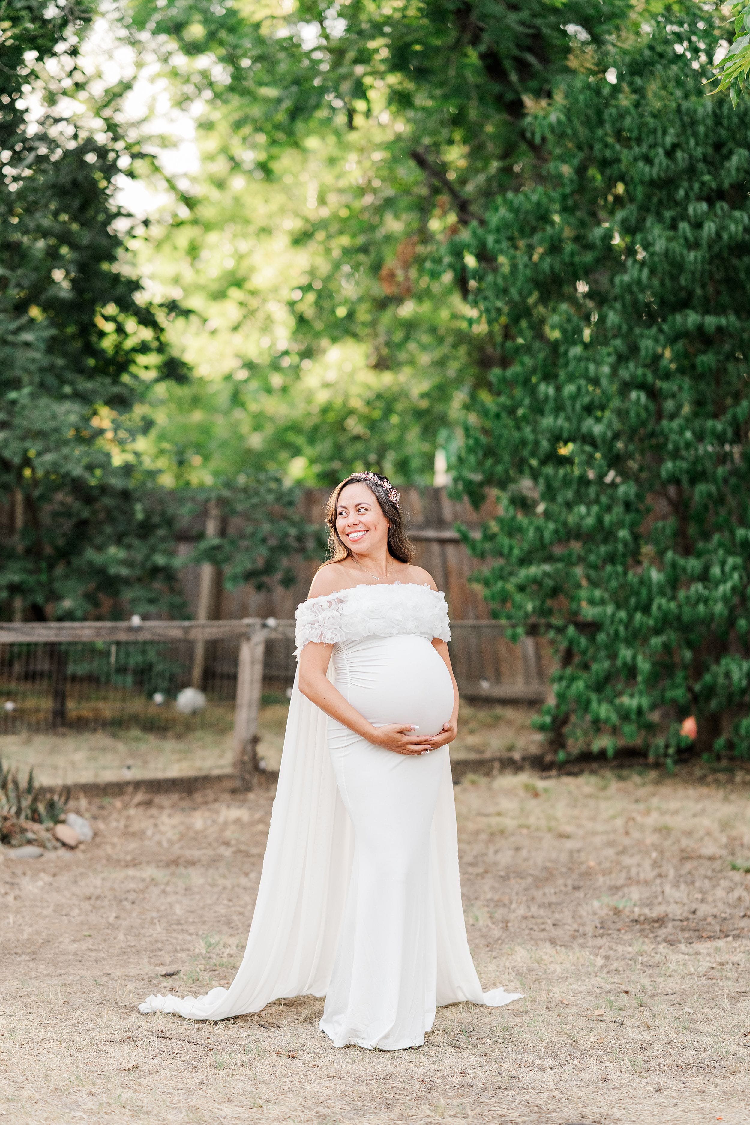 Pregnant bride in an off-the-shoulder white gown cradles her belly in a leafy garden, smiling warmly in a serene, joyful scene.