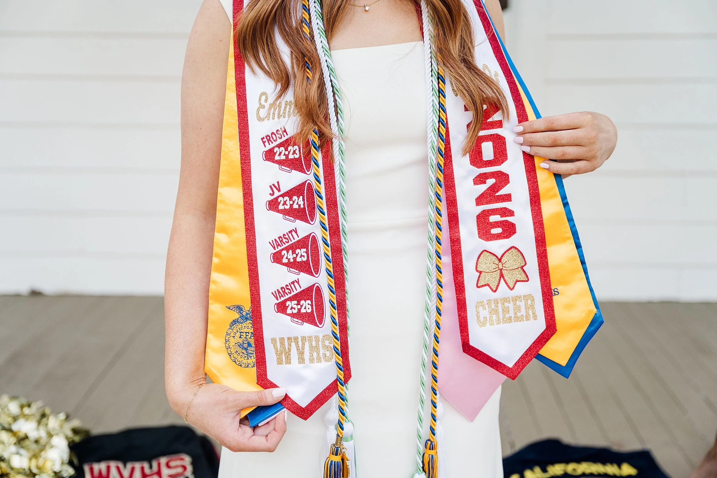 A person in a white dress wears multiple graduation stoles and cords, featuring text like "2026 Cheer" and "Frosh 22-23." The setting is light and celebratory, with cheerleading pom-poms blurred in the background.