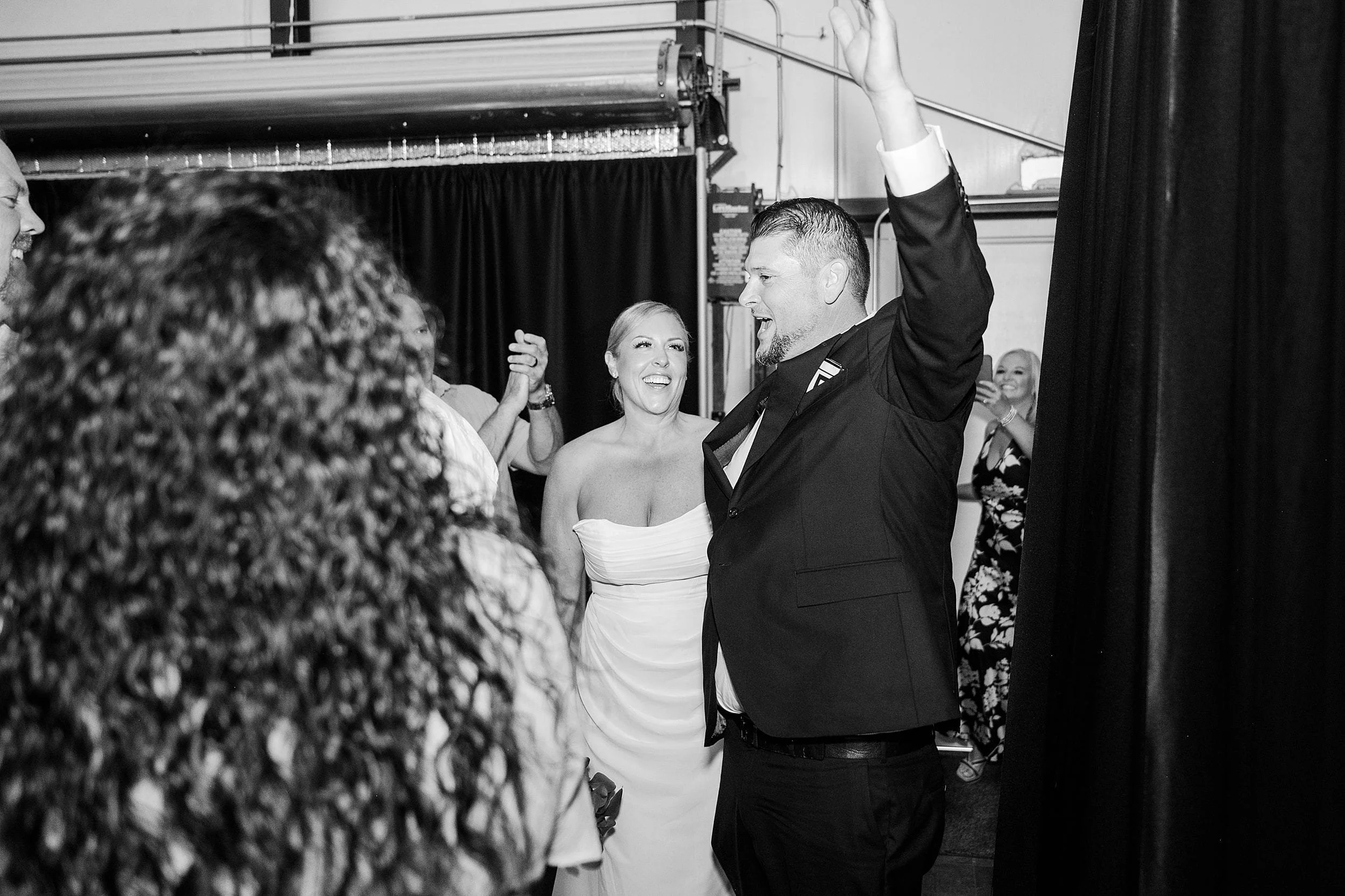 A joyful black and white wedding scene with a bride in a strapless gown and a man in a suit celebrating among guests, exuding happiness and excitement.