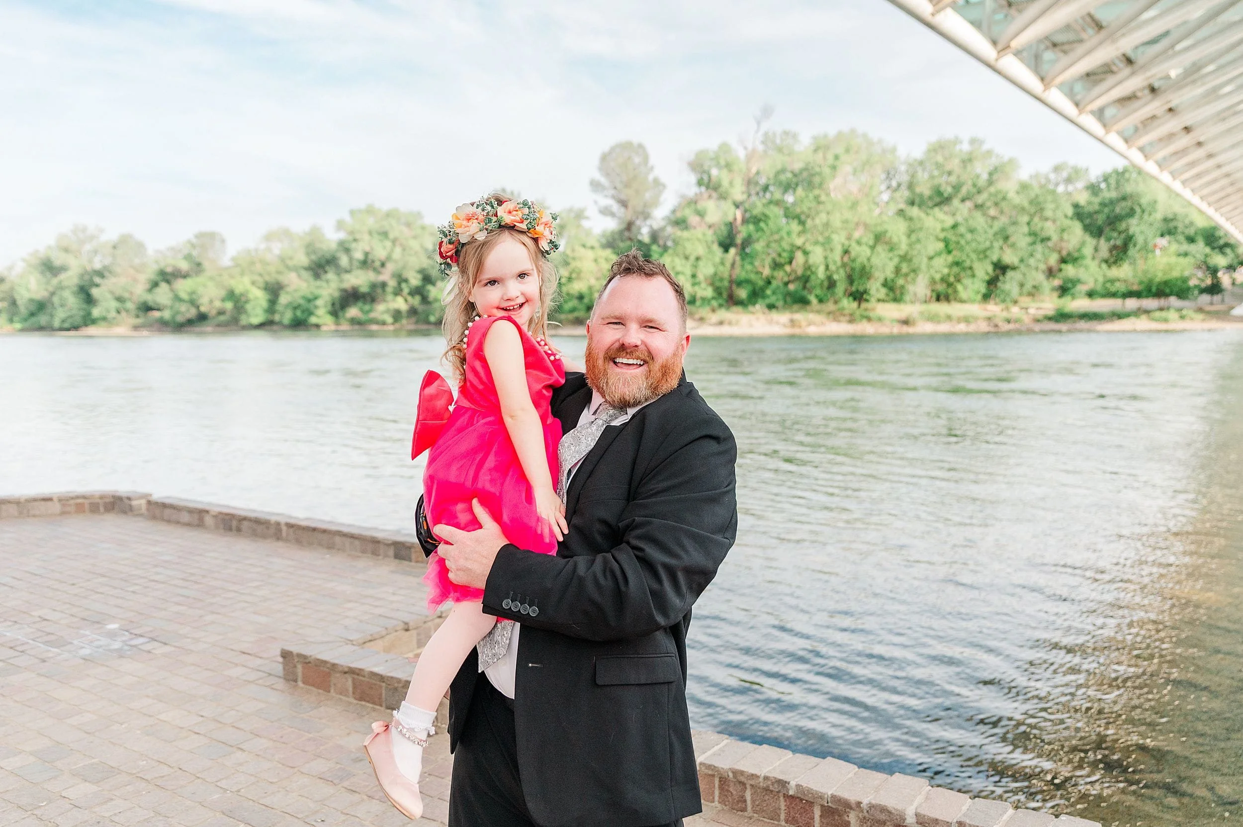 A man in a black suit holds a smiling girl in a red dress and flower crown by a river. They seem joyful. Trees and a bridge are in the background.