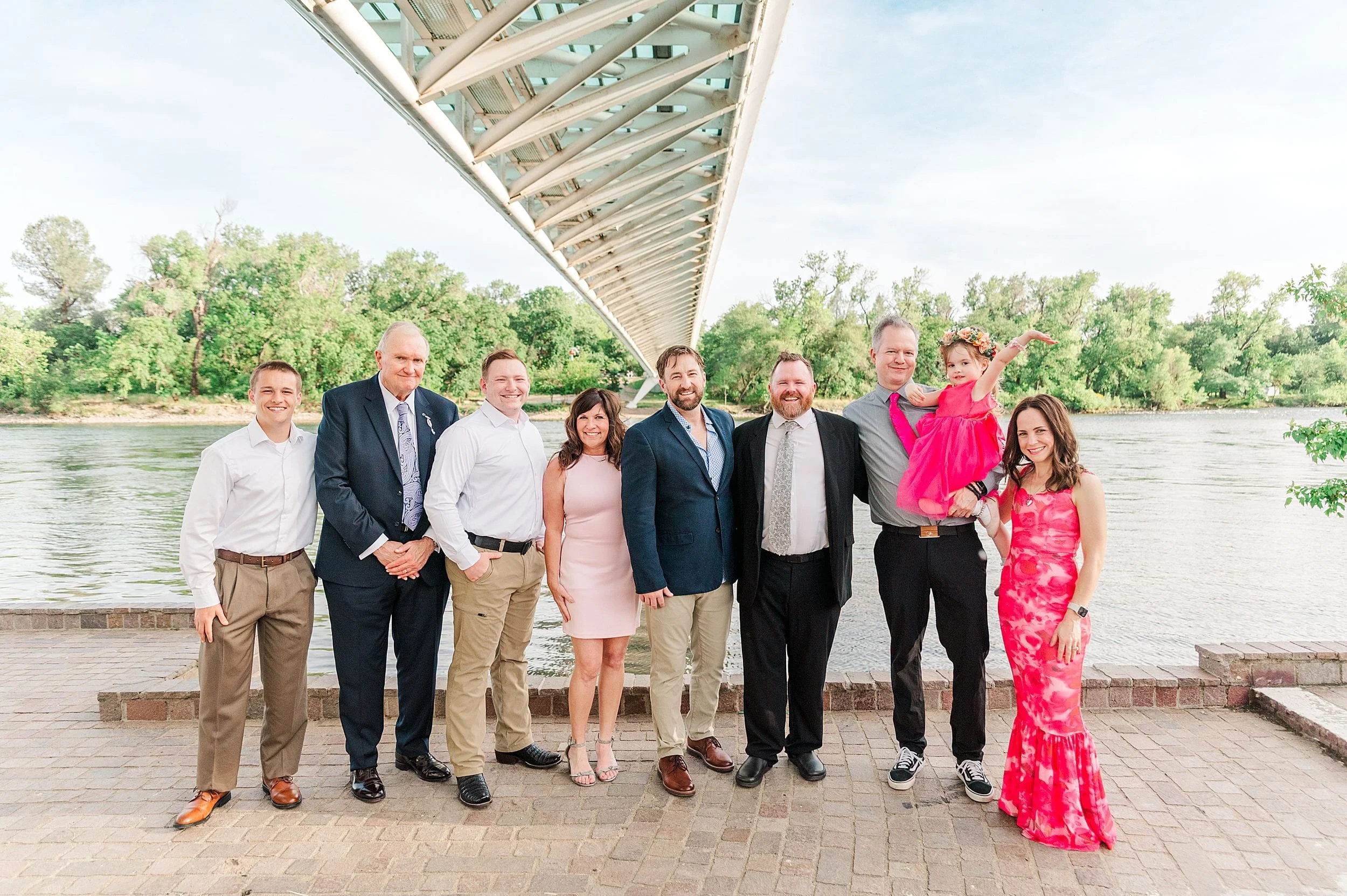 A group of eight adults and one child pose happily under a modern bridge with a river and trees in the background. The mood is celebratory and cheerful.