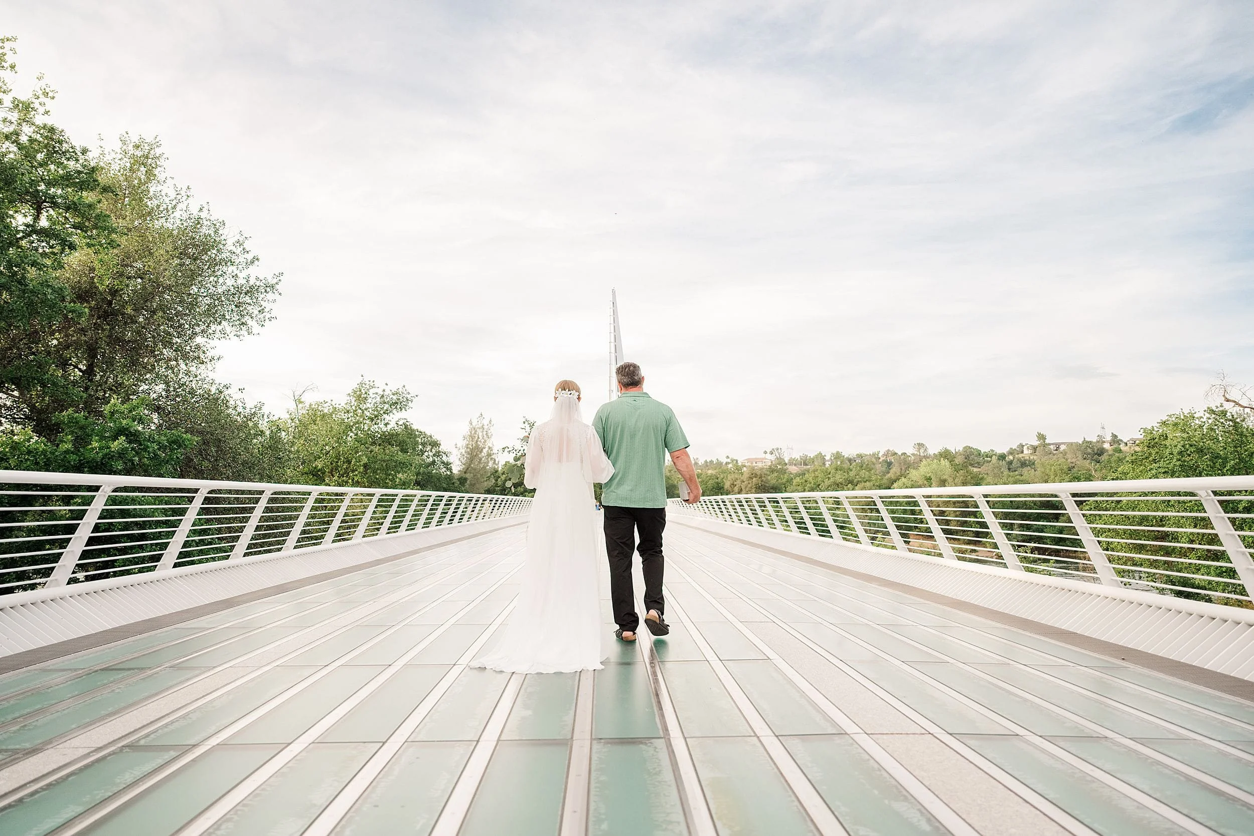 A bride in white and her father in a green shirt walk hand-in-hand across a modern bridge, surrounded by lush greenery under a bright, cloudy sky.