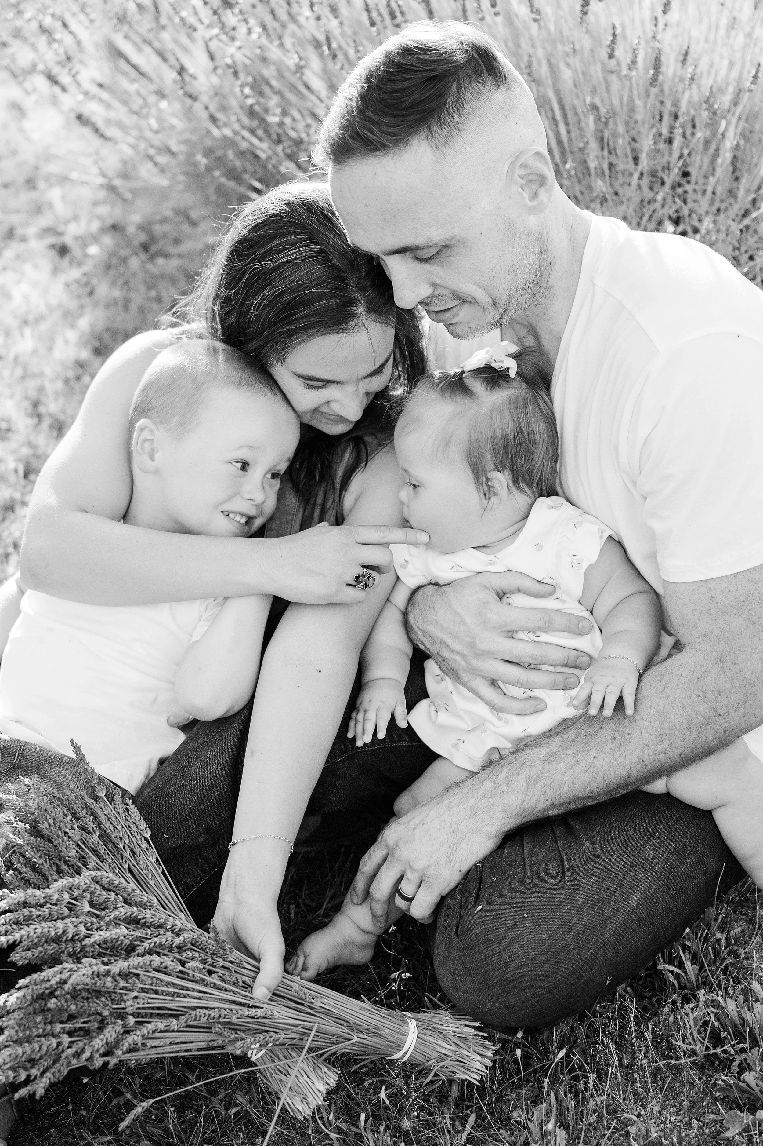 A family sits closely together outdoors, smiling warmly. The mother and father embrace their two young children, surrounded by grass and a bouquet of lavender.