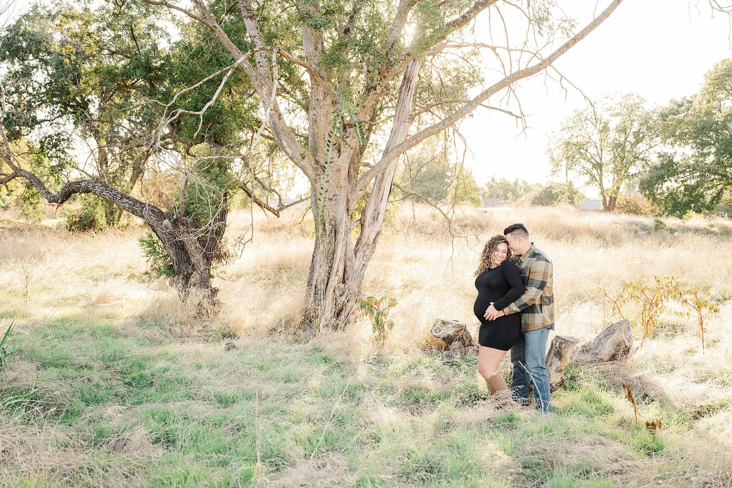 husband hugging his pregnant wife from behind standing in wide open grassy field