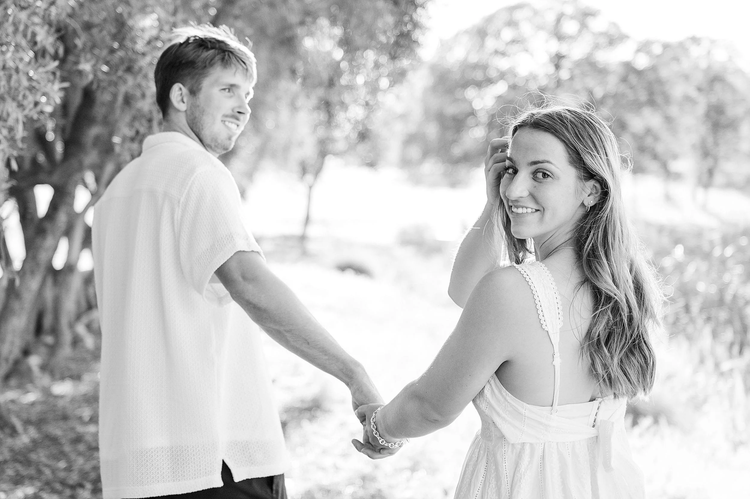 A couple holds hands, walking through a sunlit, tree-lined path. The woman looks back smiling, conveying a joyful and romantic atmosphere. Black and white.