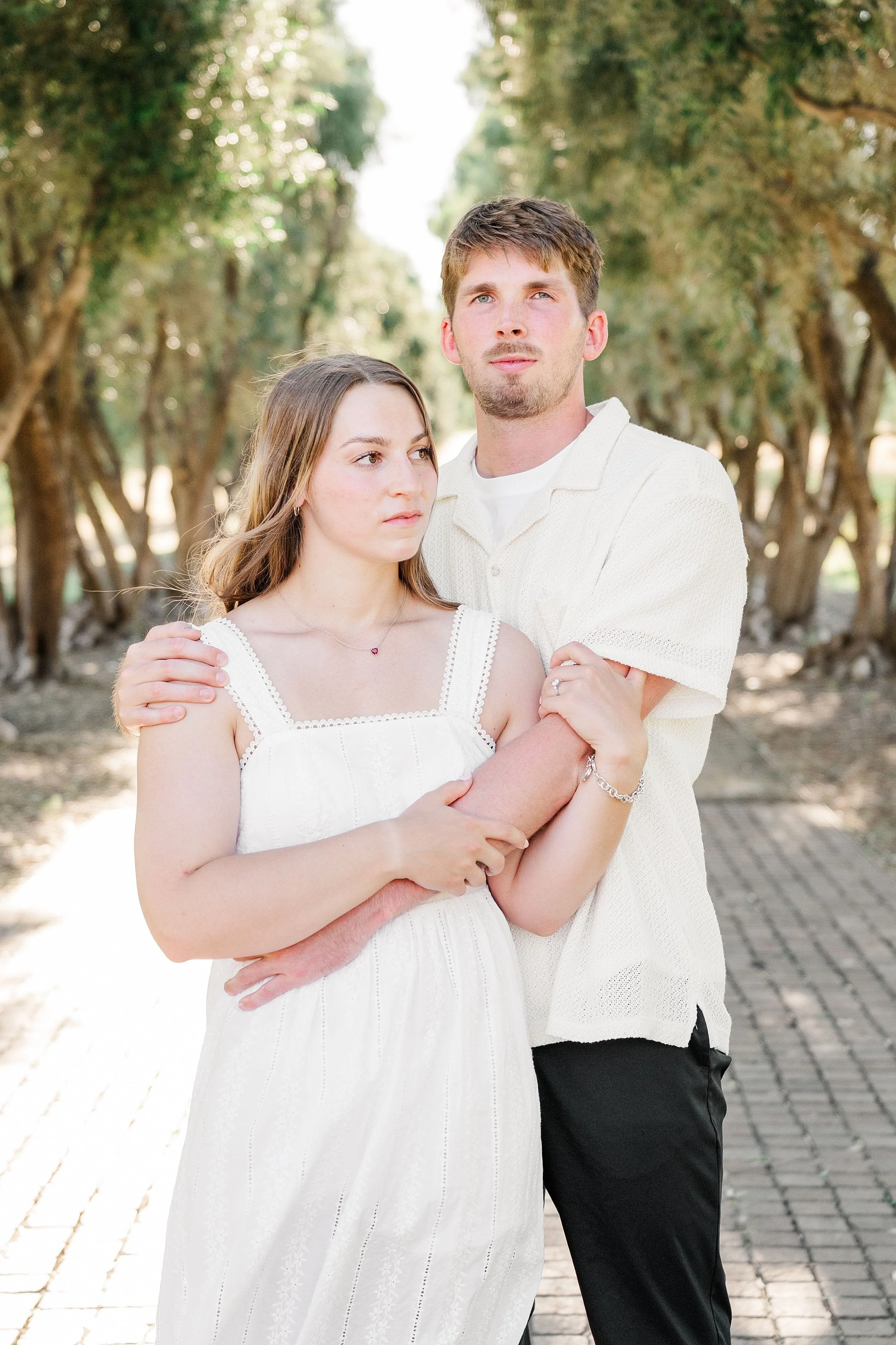 A couple standing on a sunlit path lined with trees. The woman in a white dress is embraced by the man in a light shirt. They appear calm and contemplative.