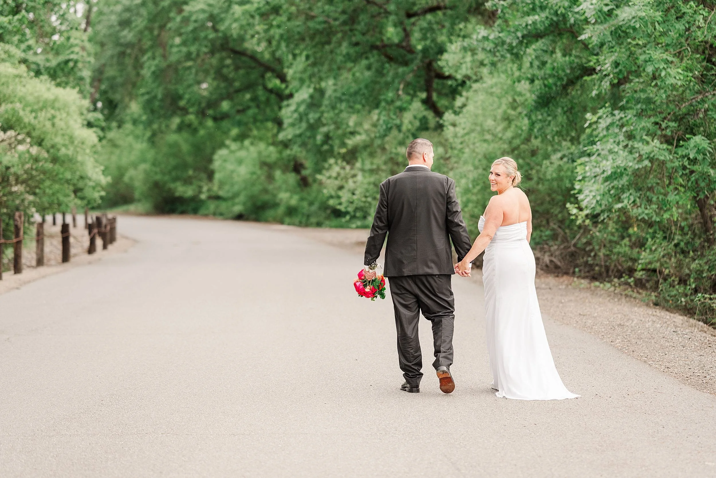 A bride in a white dress and a groom in a suit walk hand in hand down a tree-lined path. The groom holds a bouquet of red flowers, creating a romantic scene.