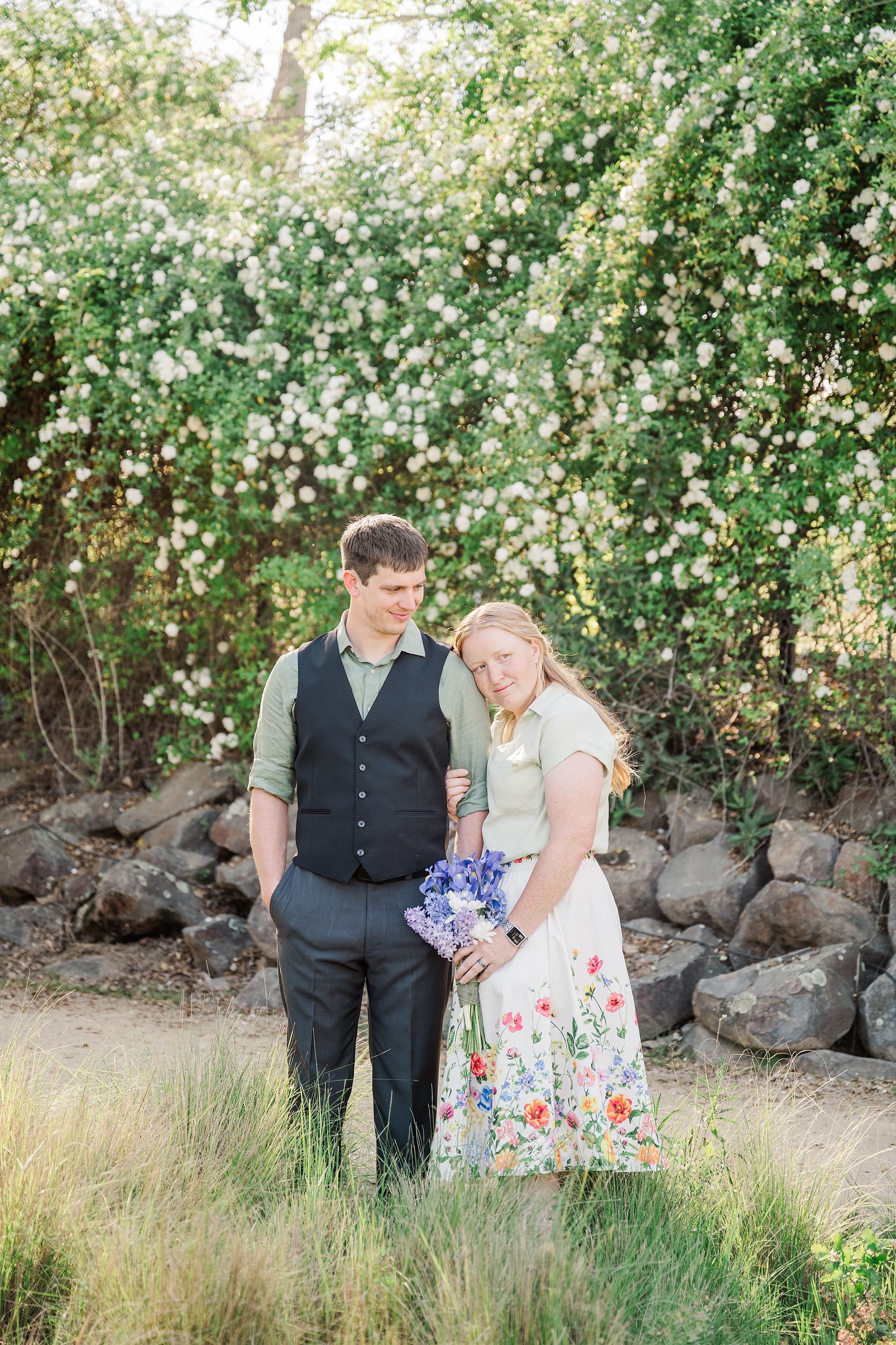 A couple stands close together in a lush garden setting, with flowering green bushes behind them. The woman, holding purple flowers, leans affectionately on the man's shoulder, creating a warm, romantic atmosphere.