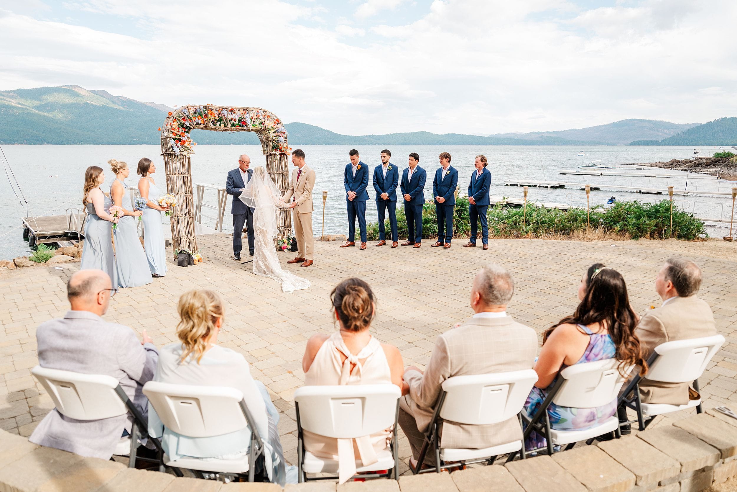 Outdoor lakeside wedding ceremony with floral arch, bride and groom, wedding party, and seated guests overlooking mountains and water.