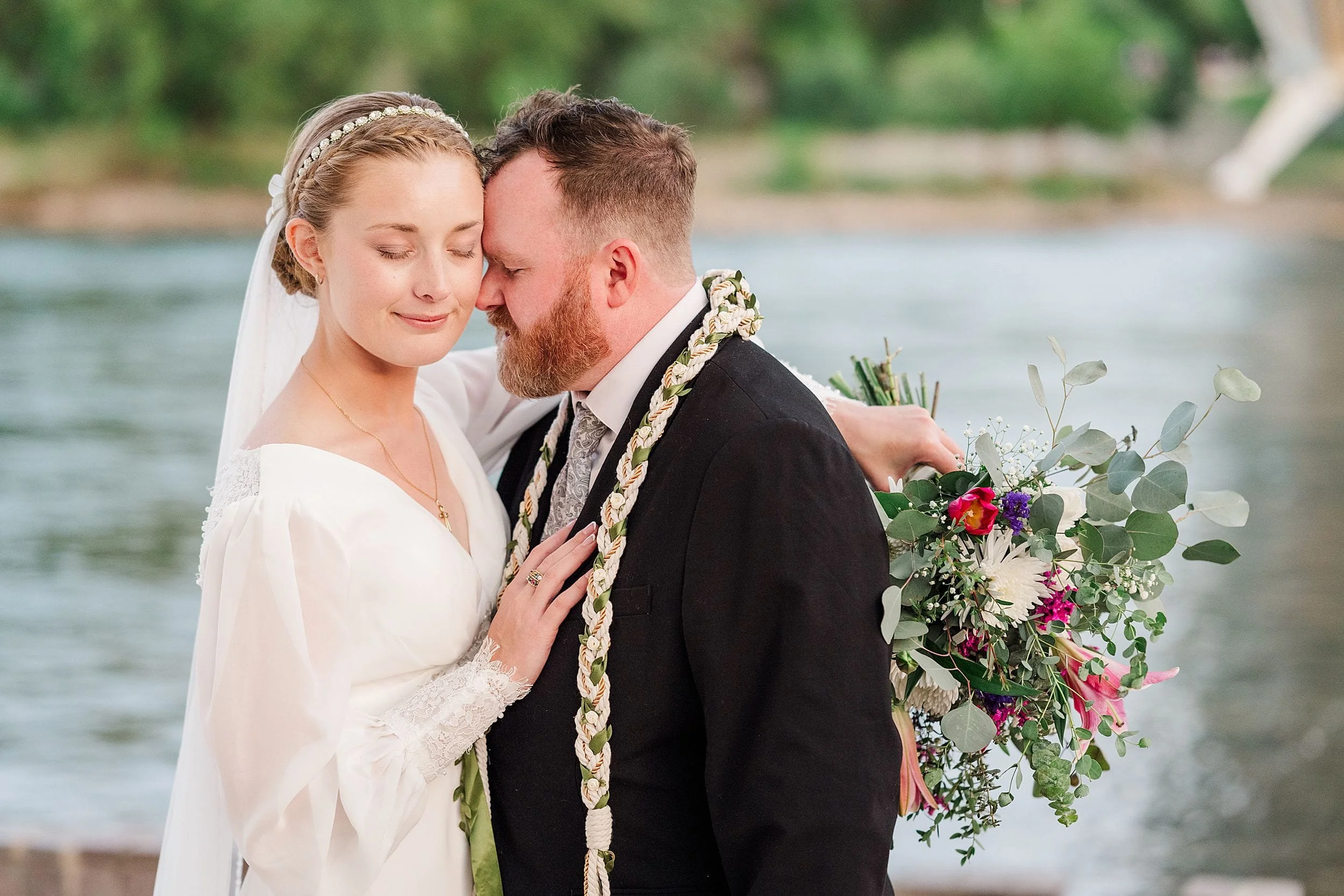 A bride and groom embrace by a river. The bride holds a bouquet of colorful flowers, both look peaceful and content. Greenery surrounds them.