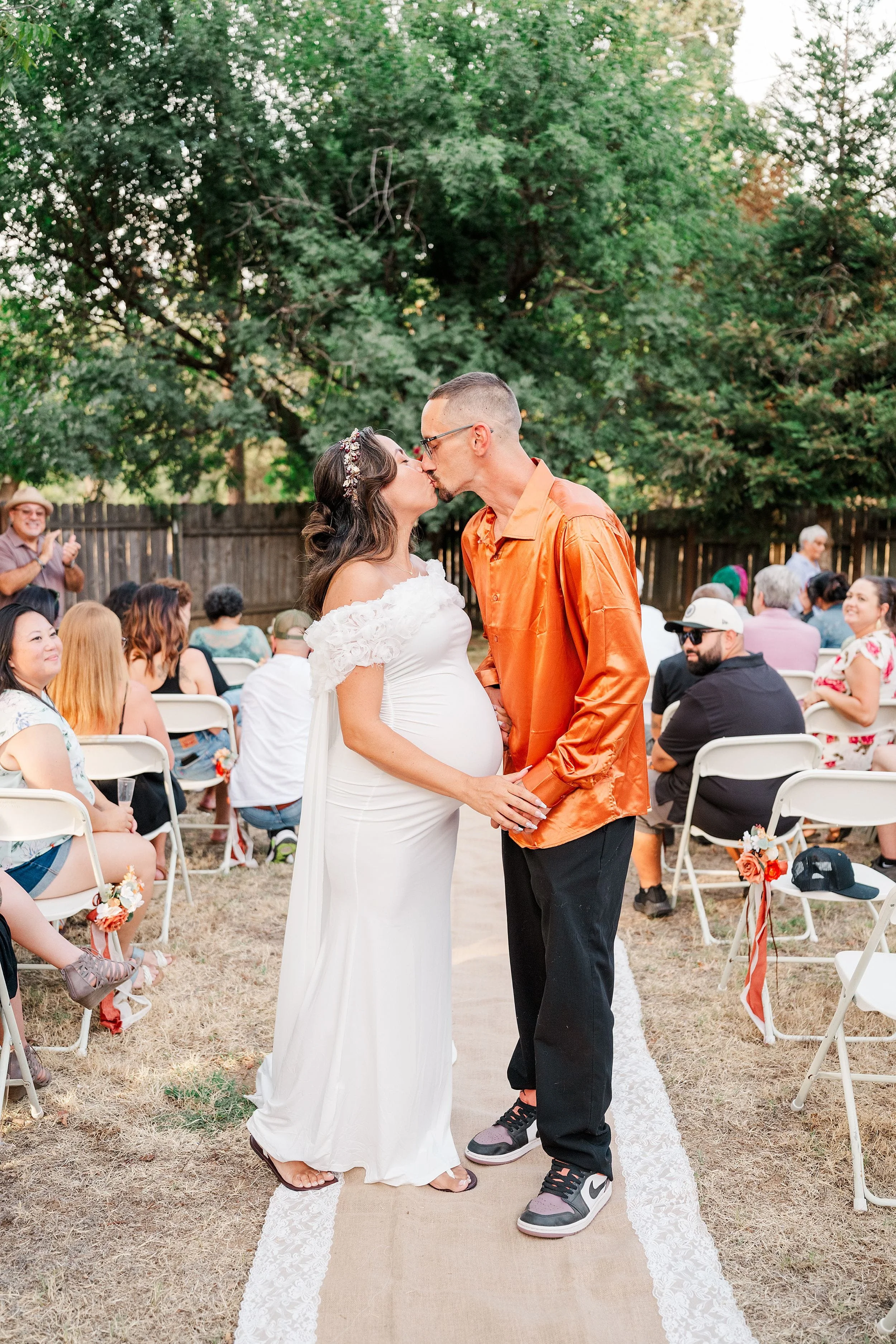 Pregnant bride in a white dress kisses her groom in an orange shirt during a joyful outdoor wedding ceremony surrounded by guests and trees.