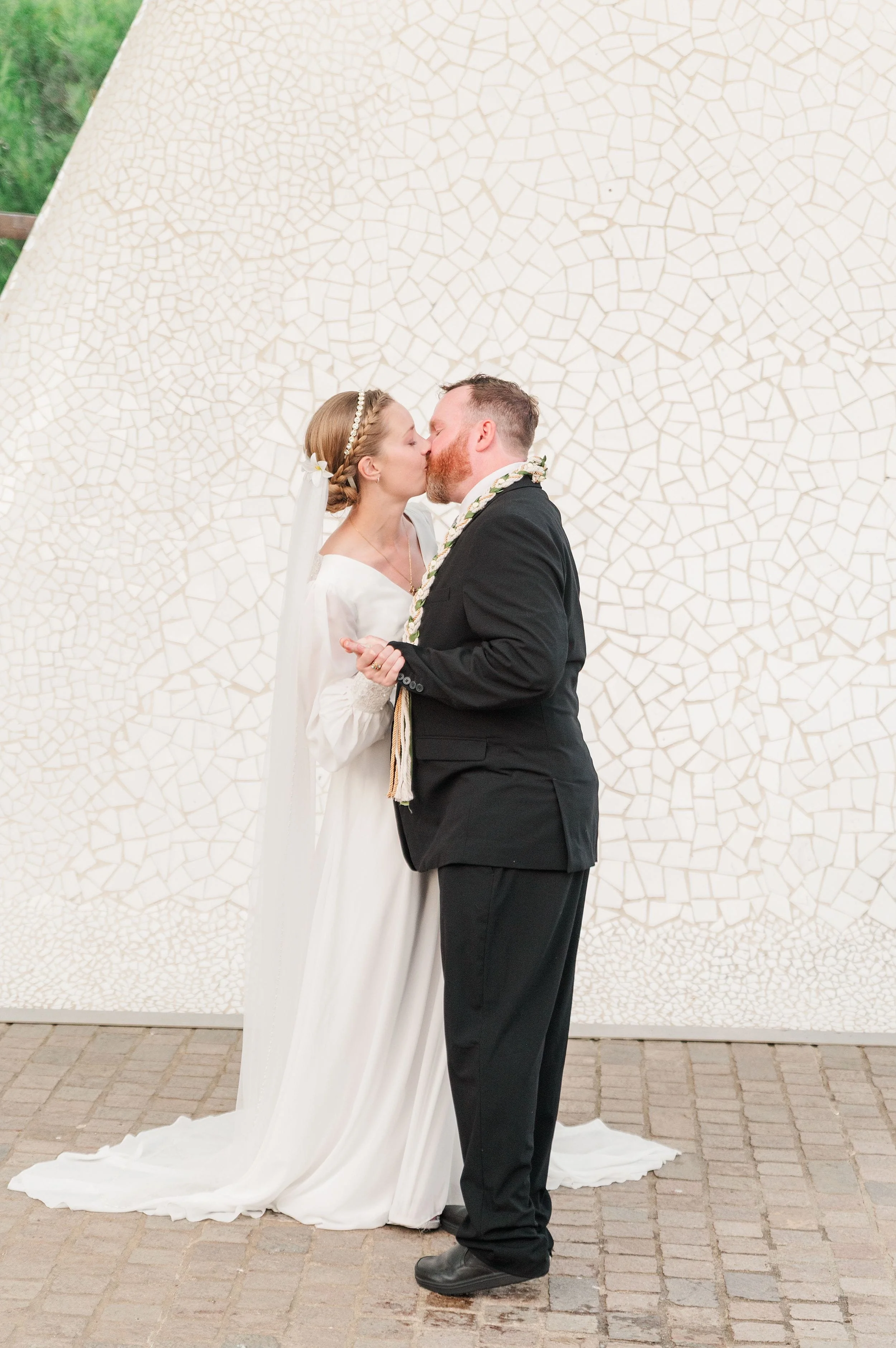 A bride and groom share a kiss in front of a white mosaic wall. She wears a long white dress and veil, while he wears a black suit, conveying joy and love.
