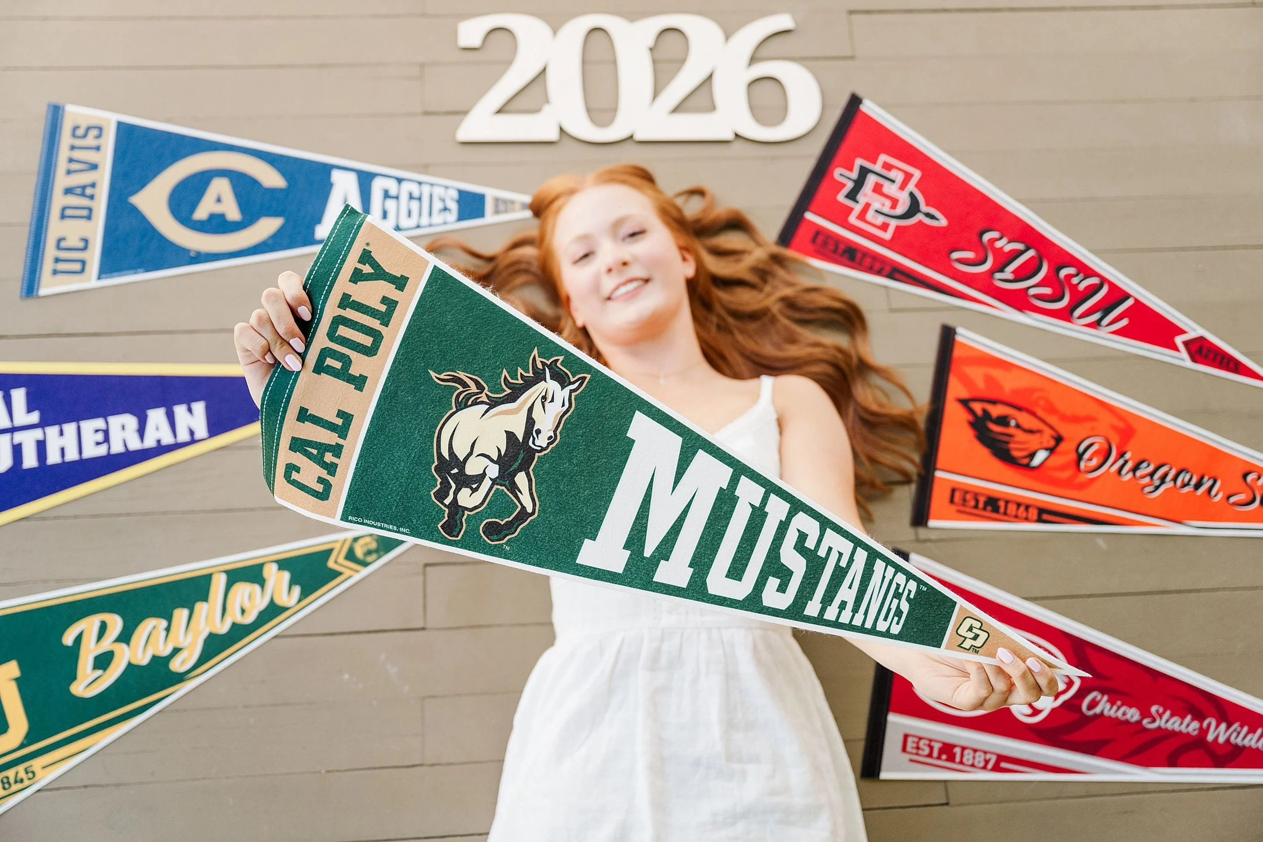 Young woman in white dress holds a green "Cal Poly Mustangs" pennant, surrounded by various college pennants on a wall. The year "2026" is above her, suggesting excitement and anticipation.