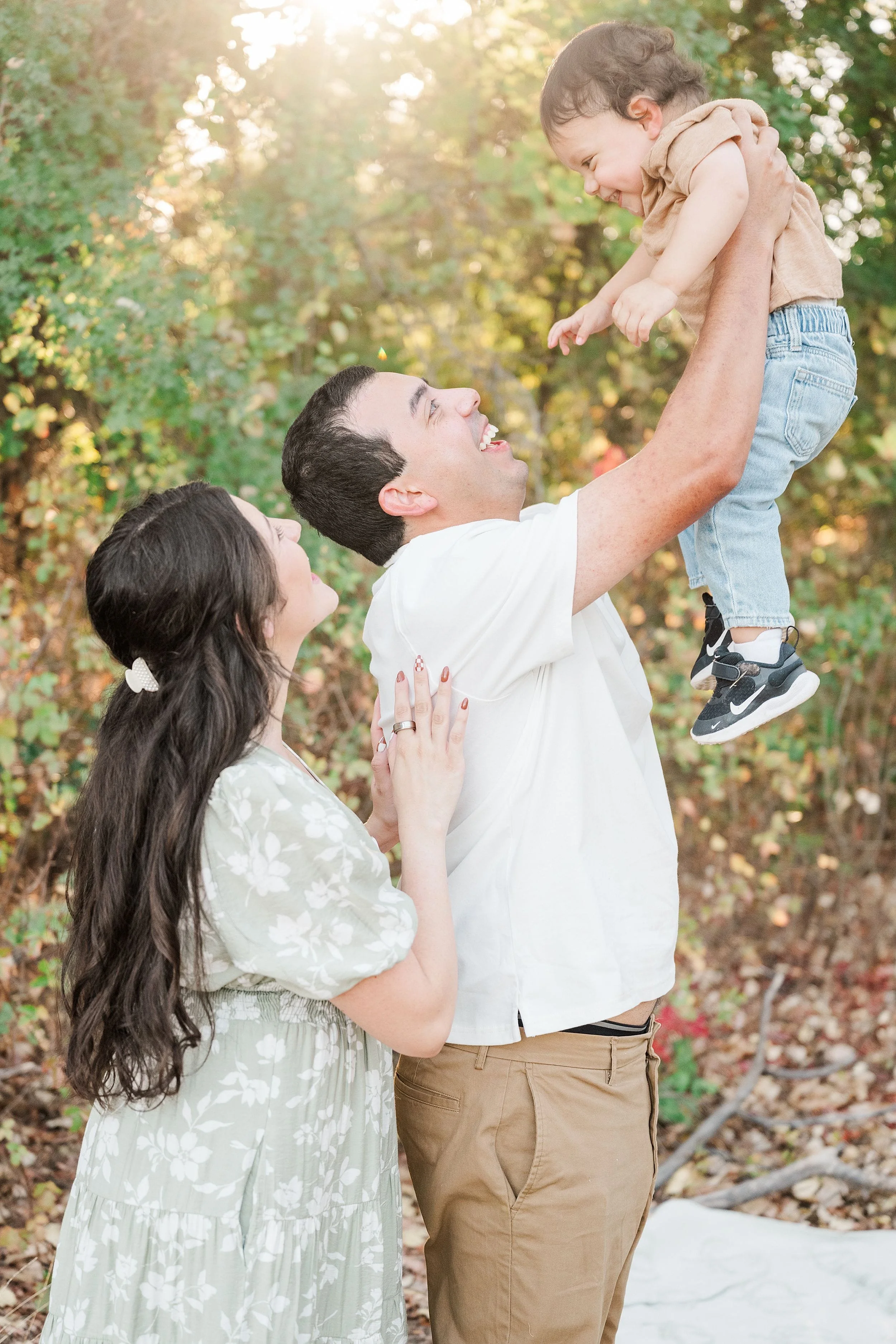 A smiling father lifts his young son overhead as her mother watches in a leafy garden, sharing a joyful, sunlit family moment.