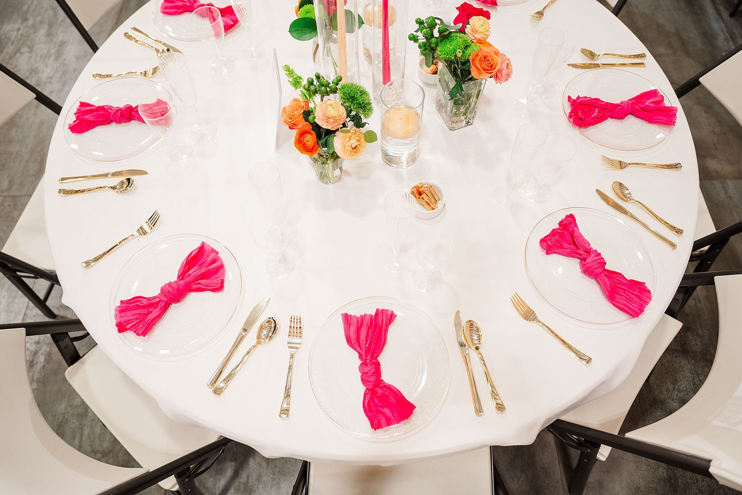Elegant round table set for an event, featuring white tablecloth, gold cutlery, pink napkins folded into bows, and vibrant floral centerpieces.