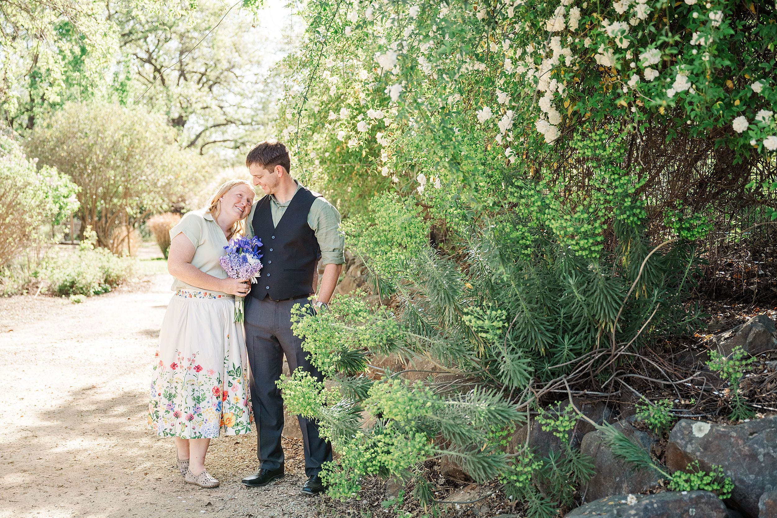 A couple stands on a sunlit garden path. The woman, in a floral skirt, holds purple flowers, leaning affectionately on the man in a vest. Blooming greenery surrounds them.