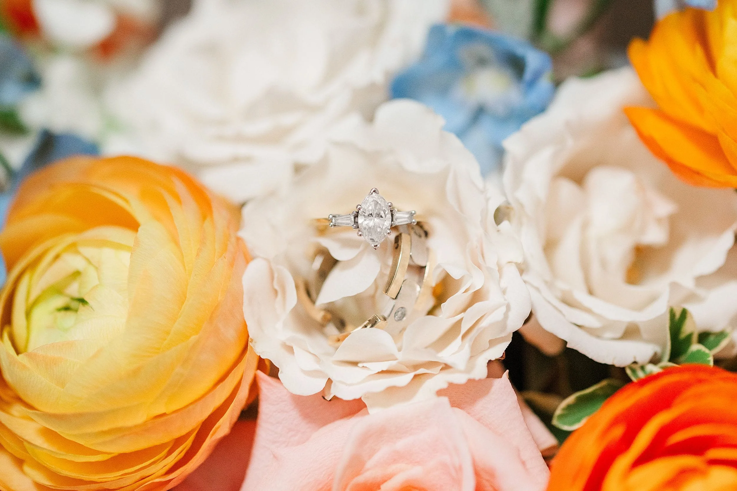 Close-up of a diamond engagement ring nestled among white, pink, orange, and blue flowers, creating a romantic, celebratory mood.