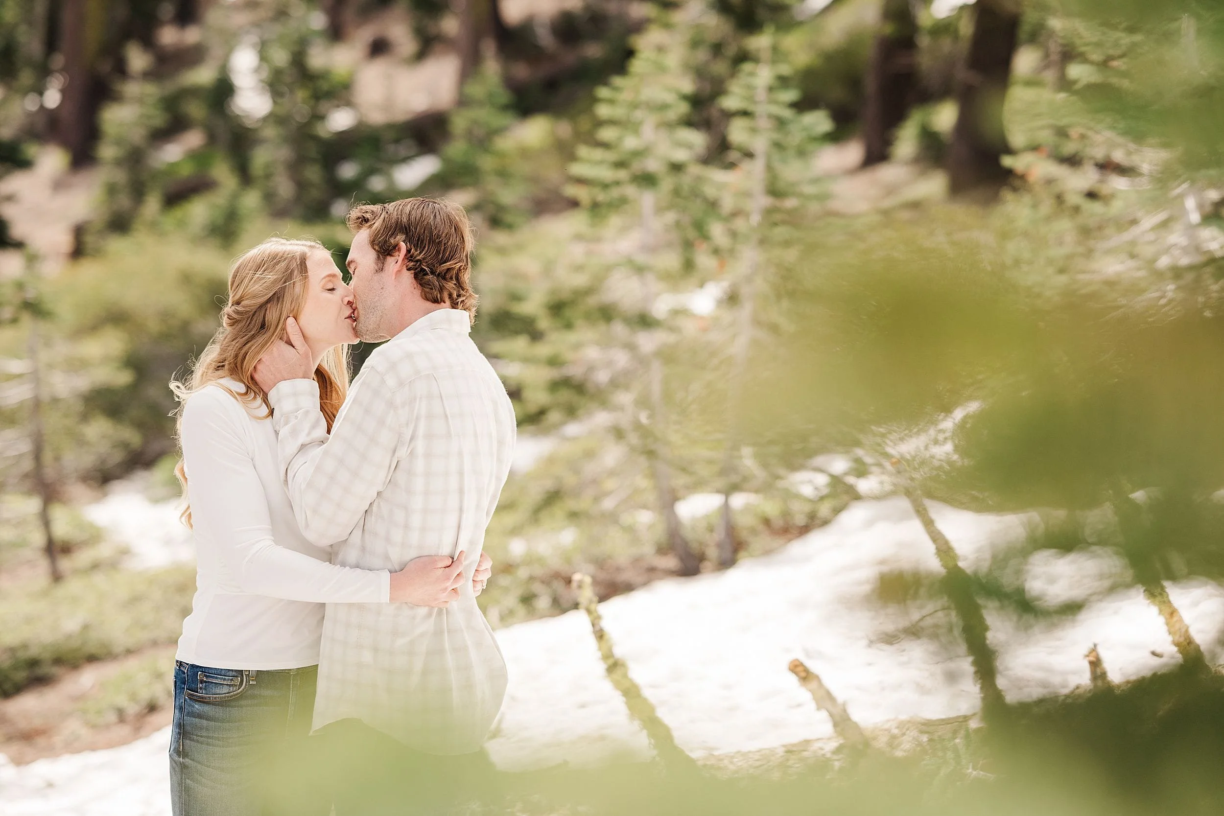 A couple kisses in a sunlit forest clearing, surrounded by greenery and patches of snow. They wear casual, light-colored clothing, conveying warmth and affection.