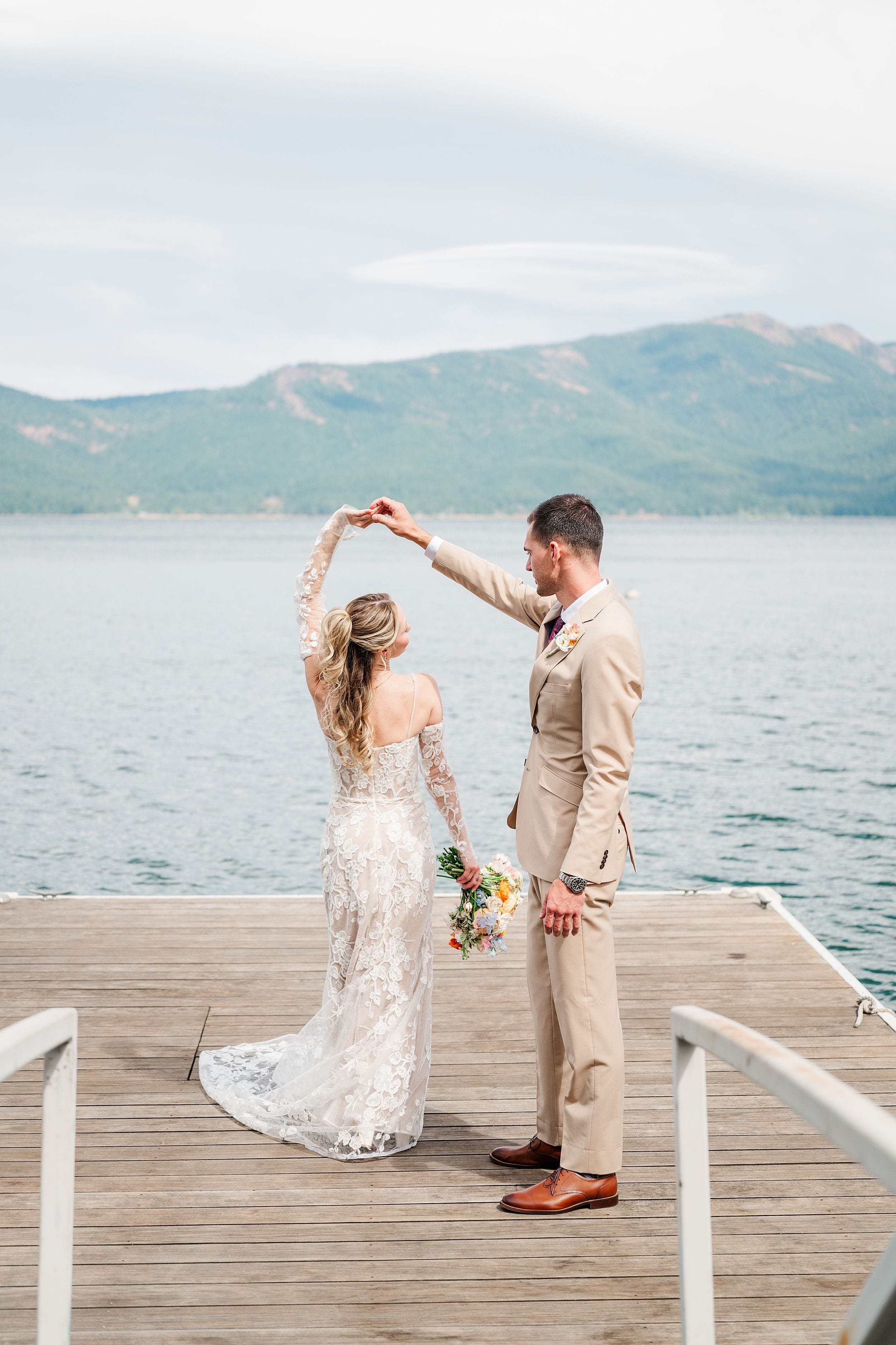 Newlywed couple shares a romantic first dance on a lakeside dock, with the bride in a lace gown and groom in a tan suit, mountains behind them.