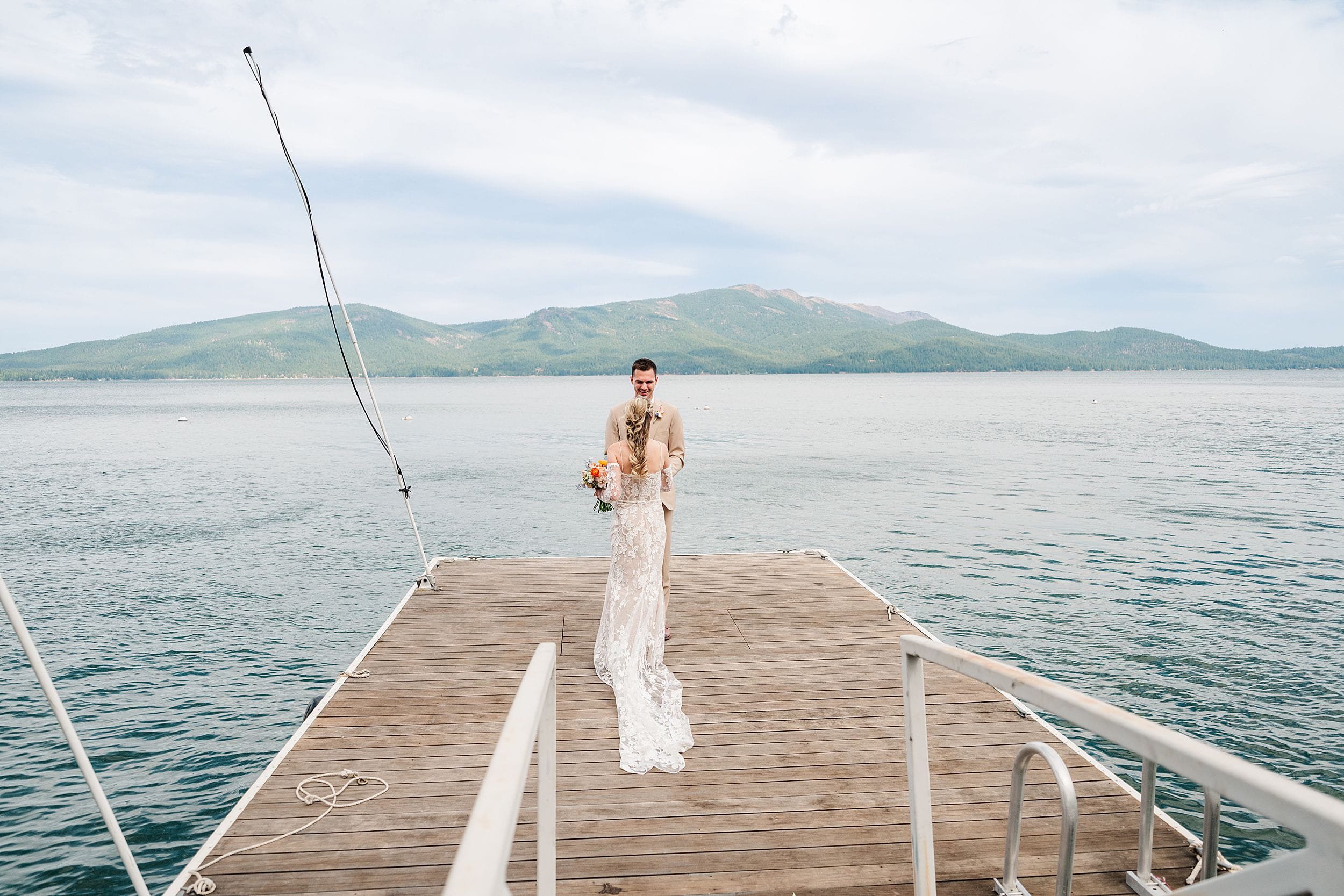 Bride in a white gown and floral bouquet stands on a wooden lakeside dock, with calm water and green mountains creating a serene, romantic scene.