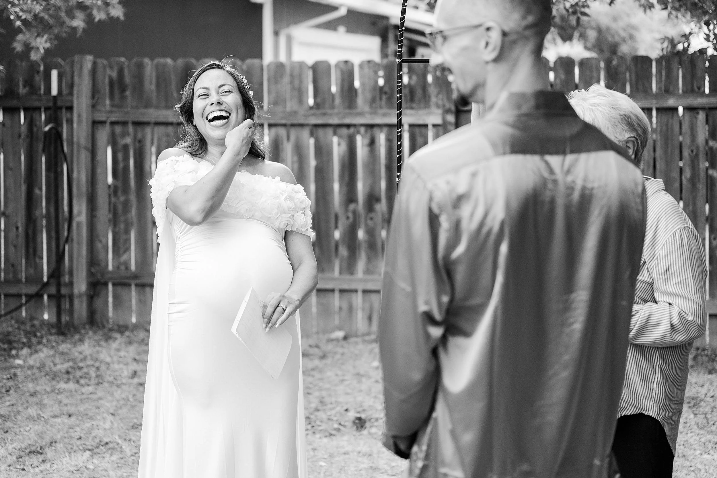 Black-and-white backyard gathering shows a joyful pregnant woman in a white dress laughing as she greets two older guests, creating a warm mood.
