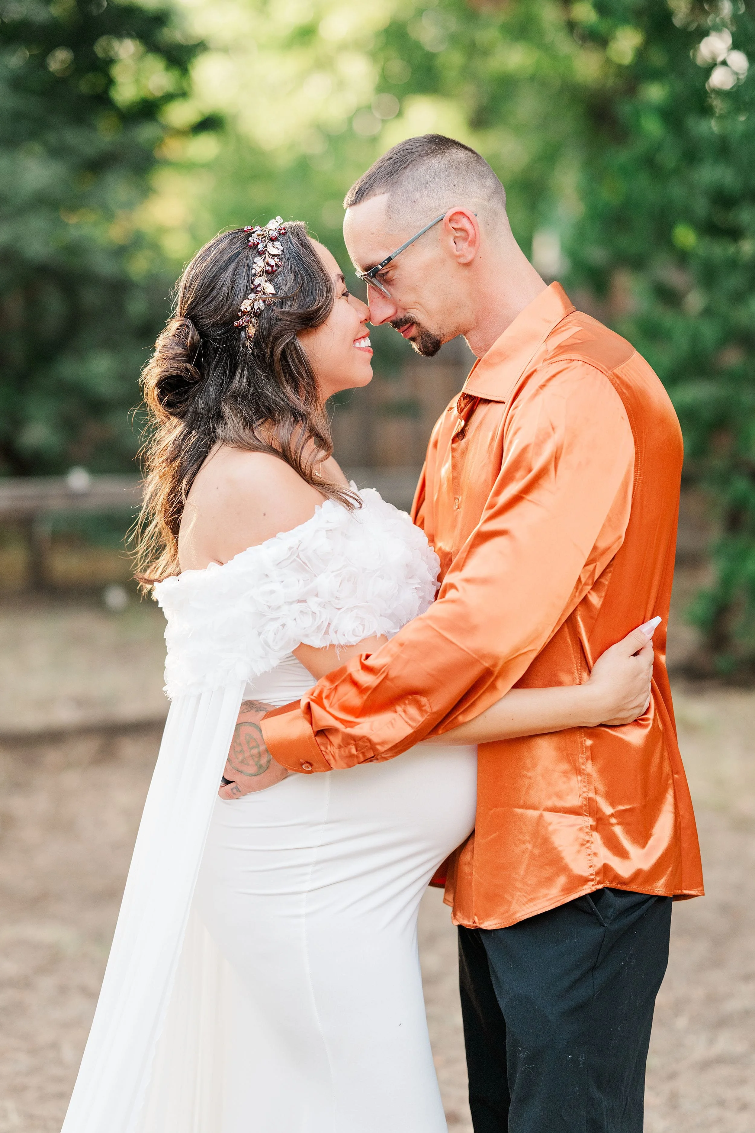 A bride in a white gown and floral hairpiece embraces a groom in a bright orange shirt outdoors, touching noses in a tender, joyful moment.