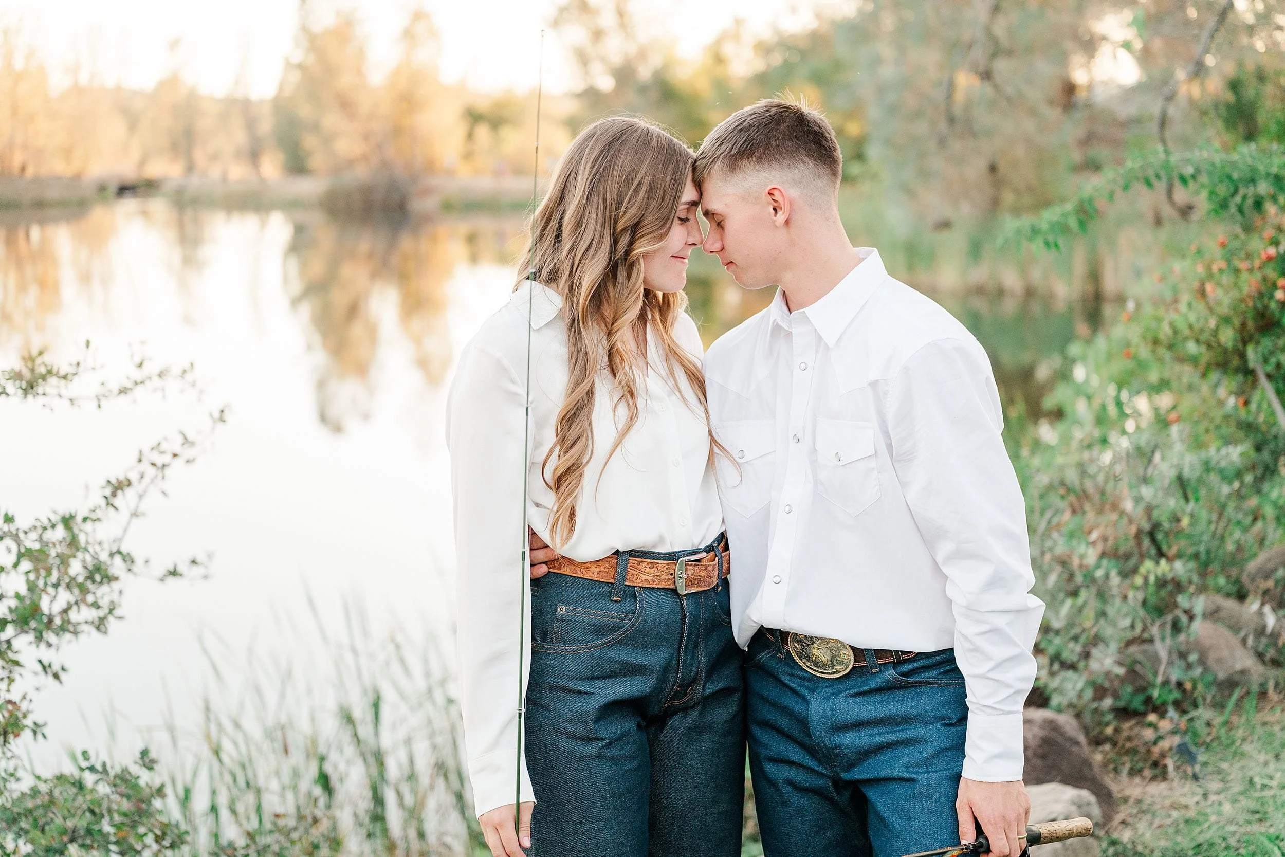A young couple in white shirts and jeans touch foreheads beside a calm lake, surrounded by autumn foliage; the soft light creates a tender, romantic mood.