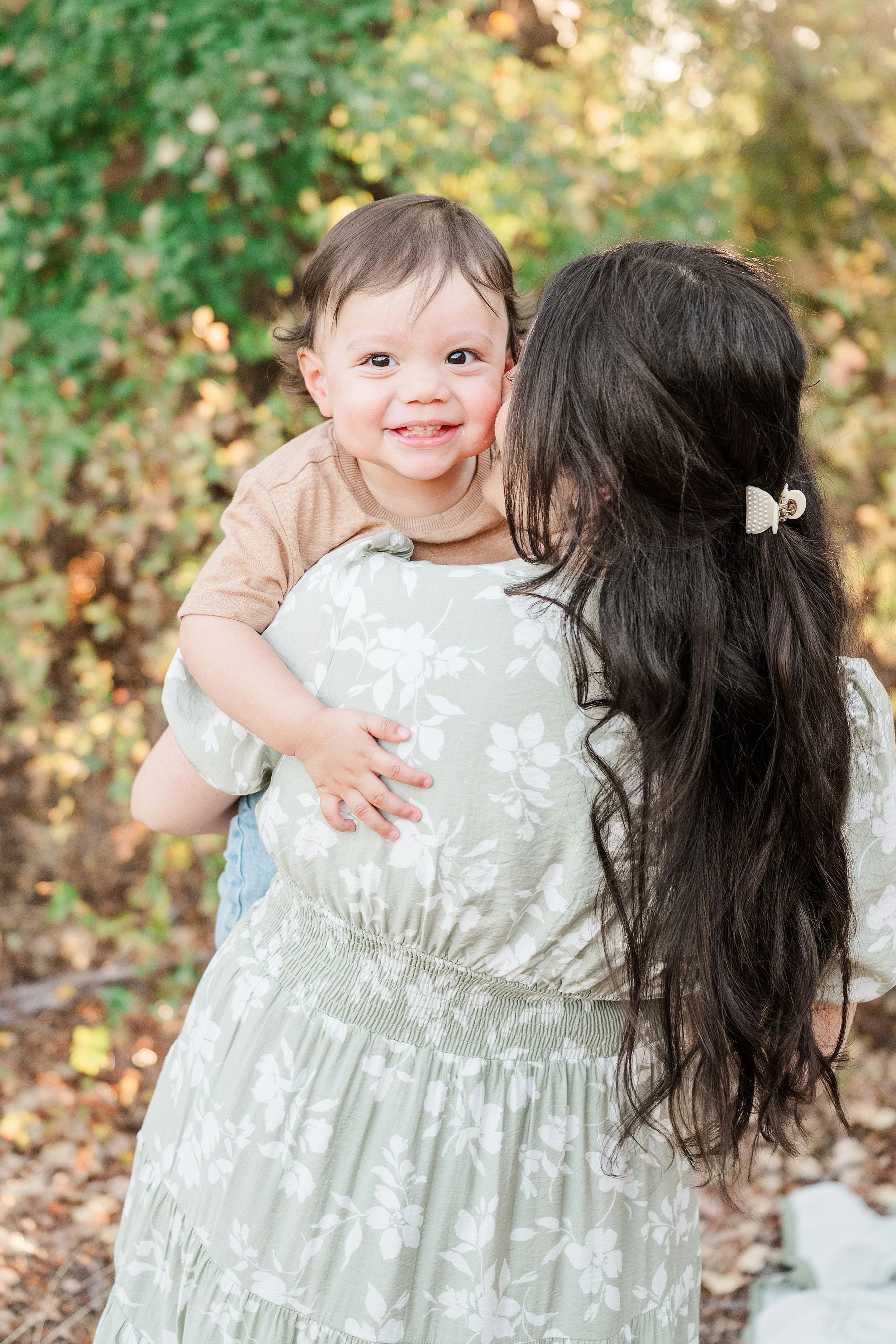 A smiling baby rests on a woman’s shoulder as she carries the child outdoors among autumn leaves, capturing a warm, tender family moment.