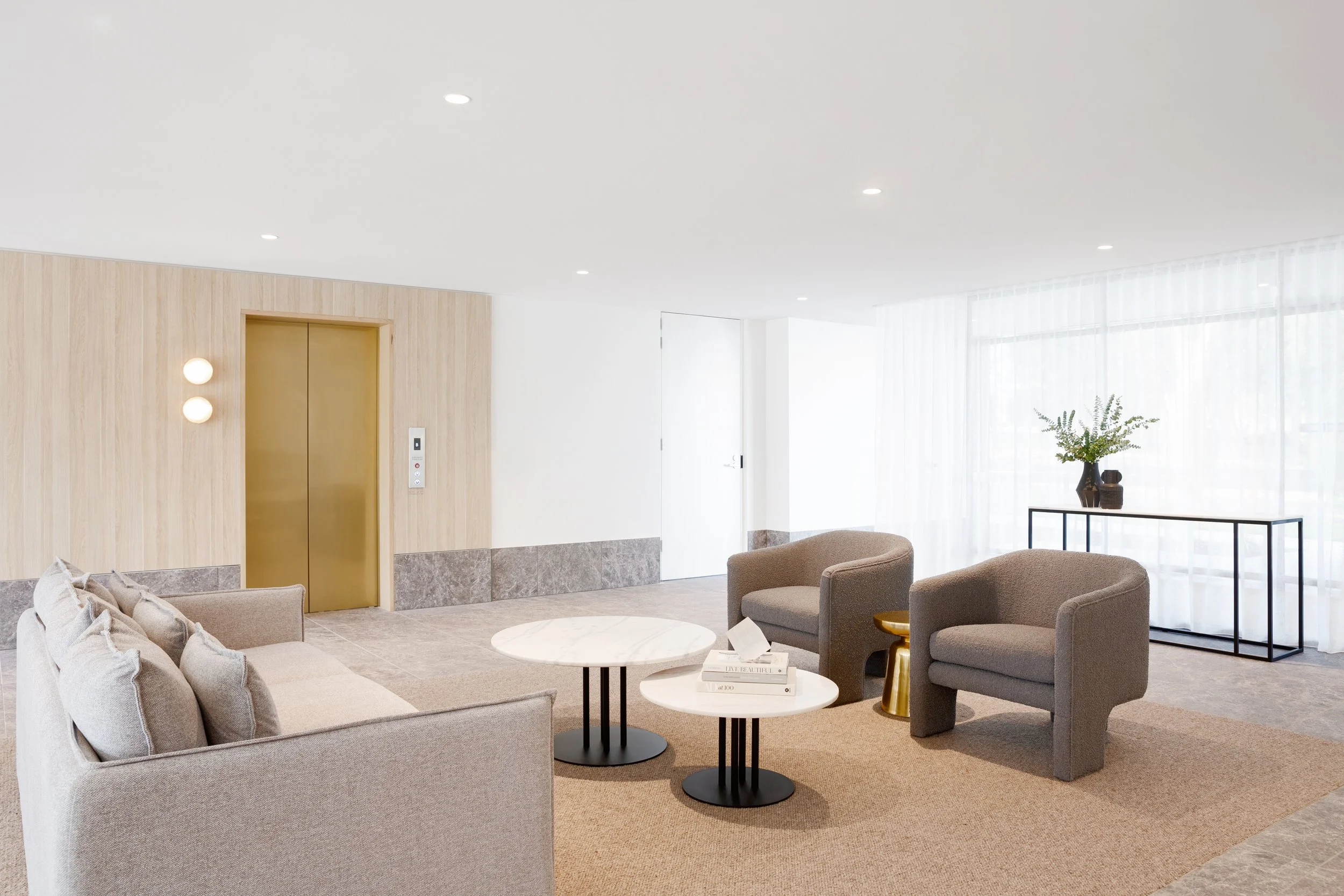 Modern hotel lobby with beige and gray furniture, round coffee tables, and large windows with sheer curtains.