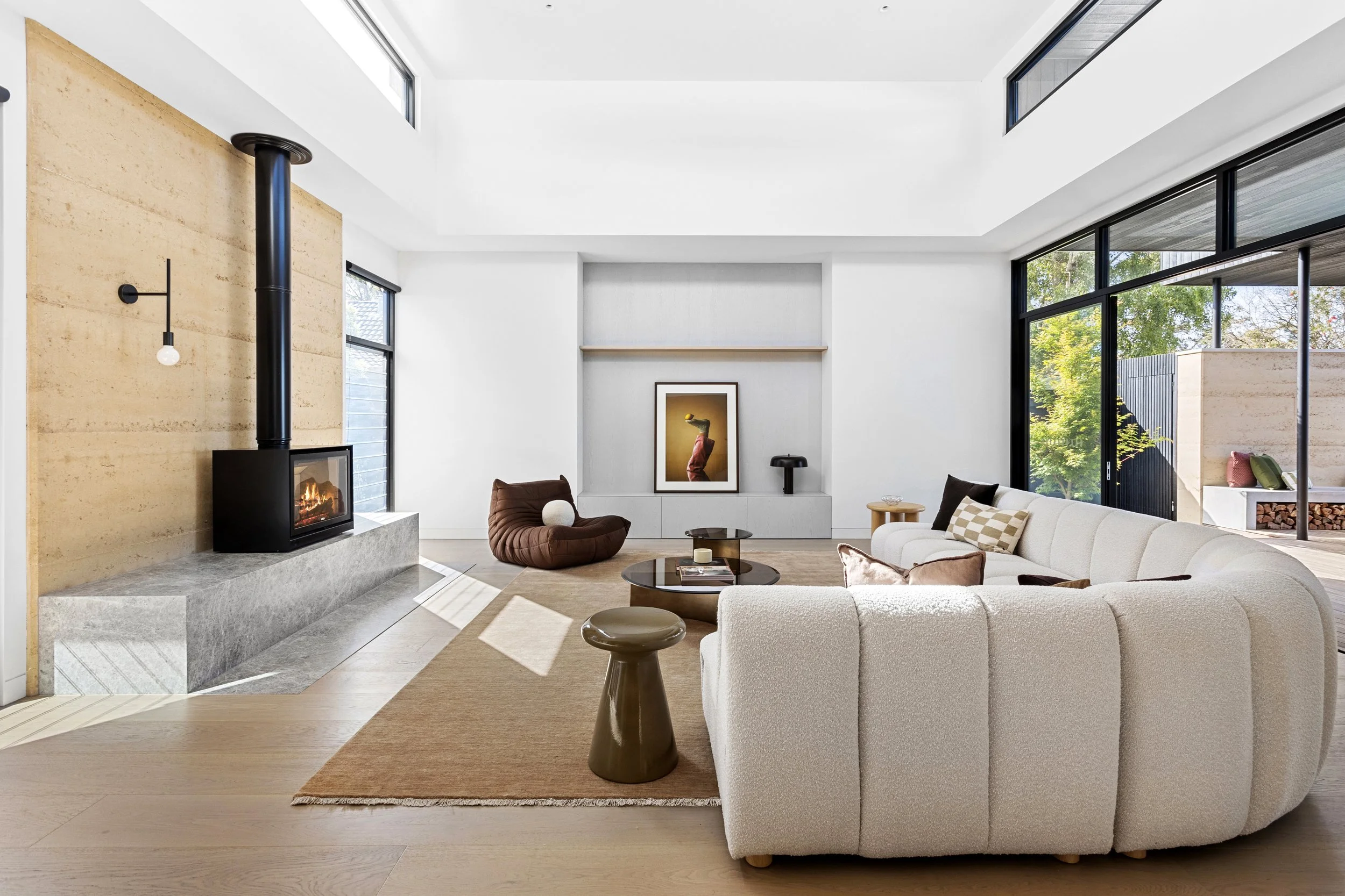 Modern living room with white walls, large windows, a fireplace, a white sofa, a brown chair, a coffee table, and framed artwork.