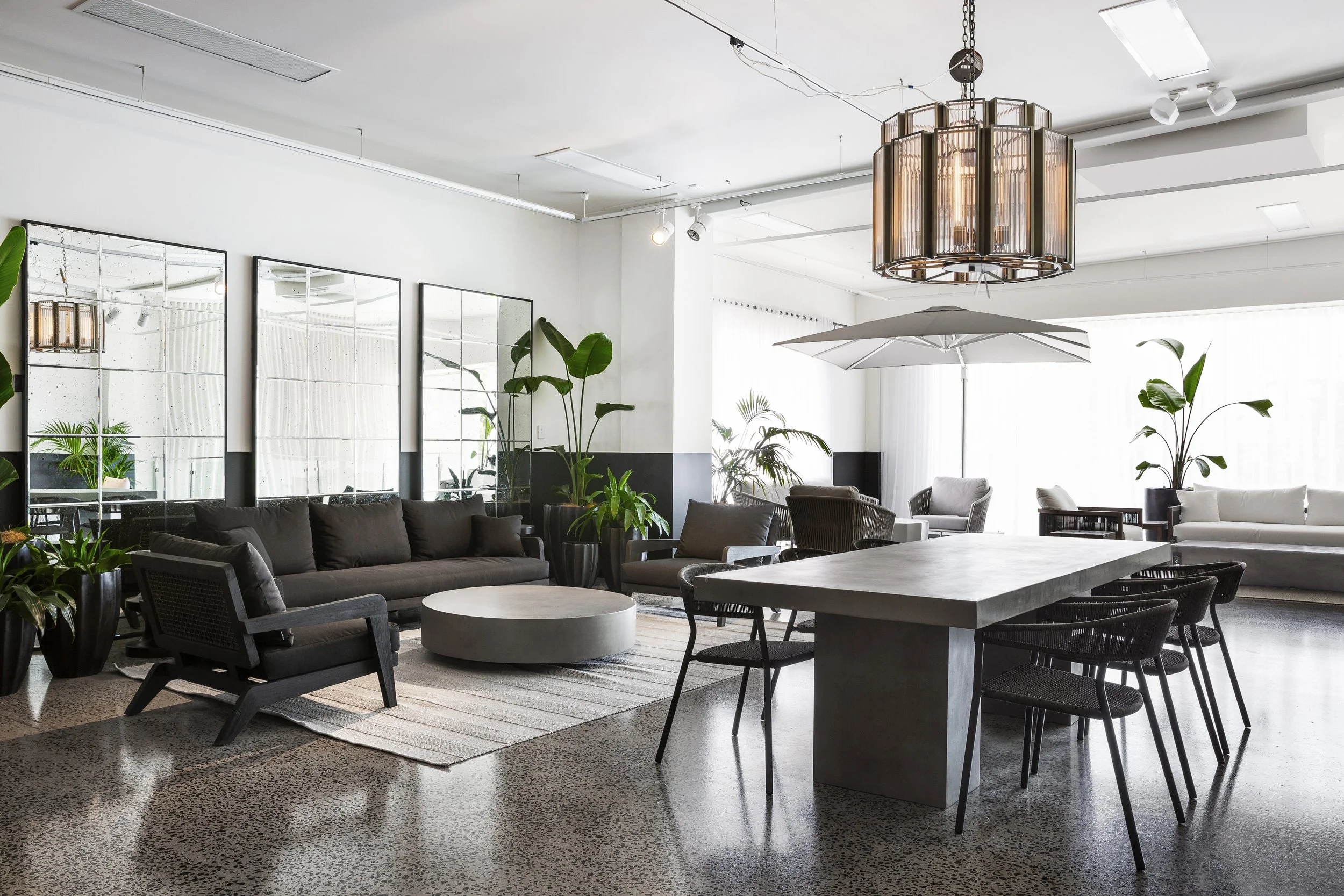 Modern indoor lounge area with black sofas, a large concrete table, white ceiling, indoor plants, and exposed ceiling lights and fixtures.