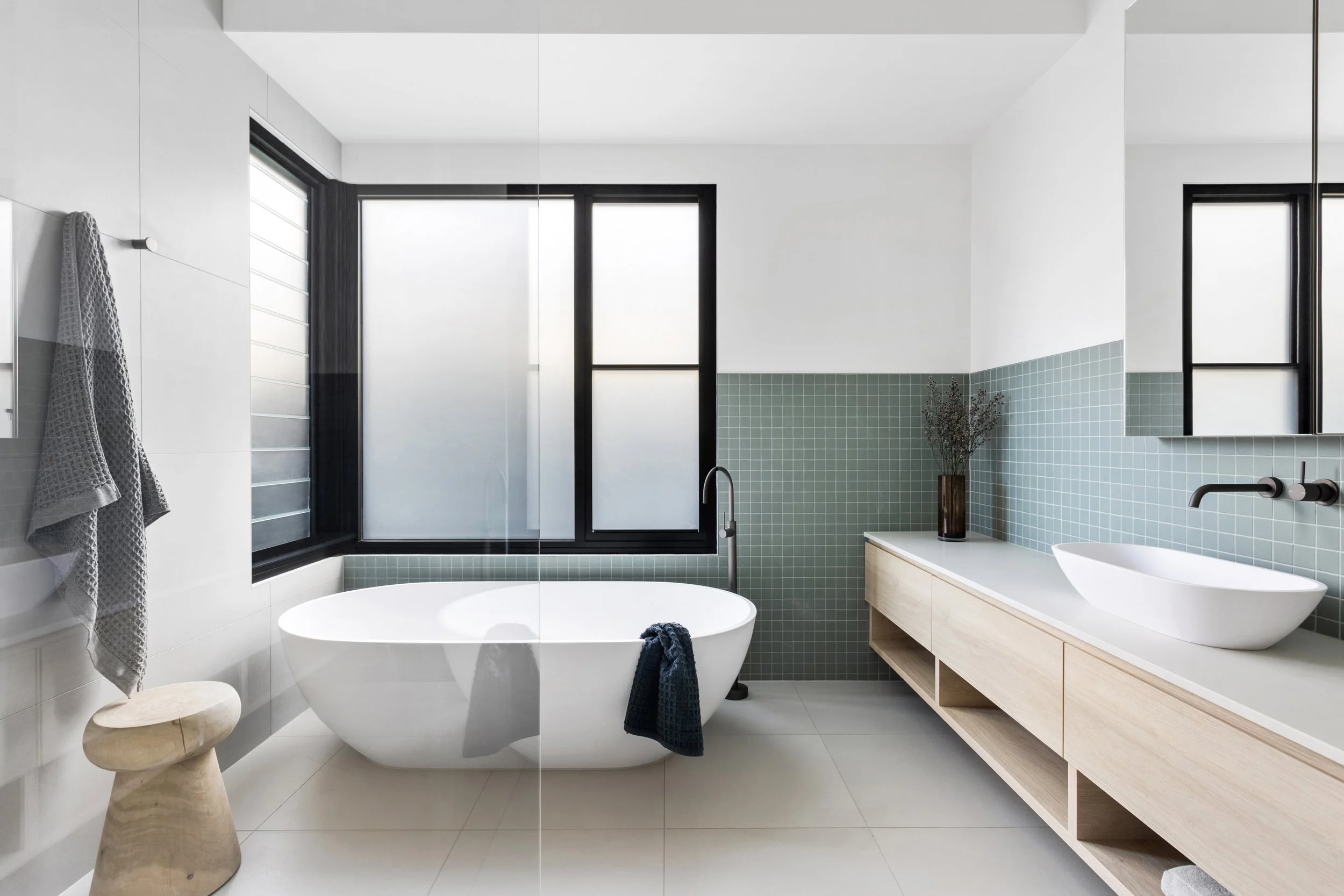 Modern bathroom with white and green tiled walls, a white bathtub, black-framed frosted window, wooden vanity with sink, and decorative dried flowers.
