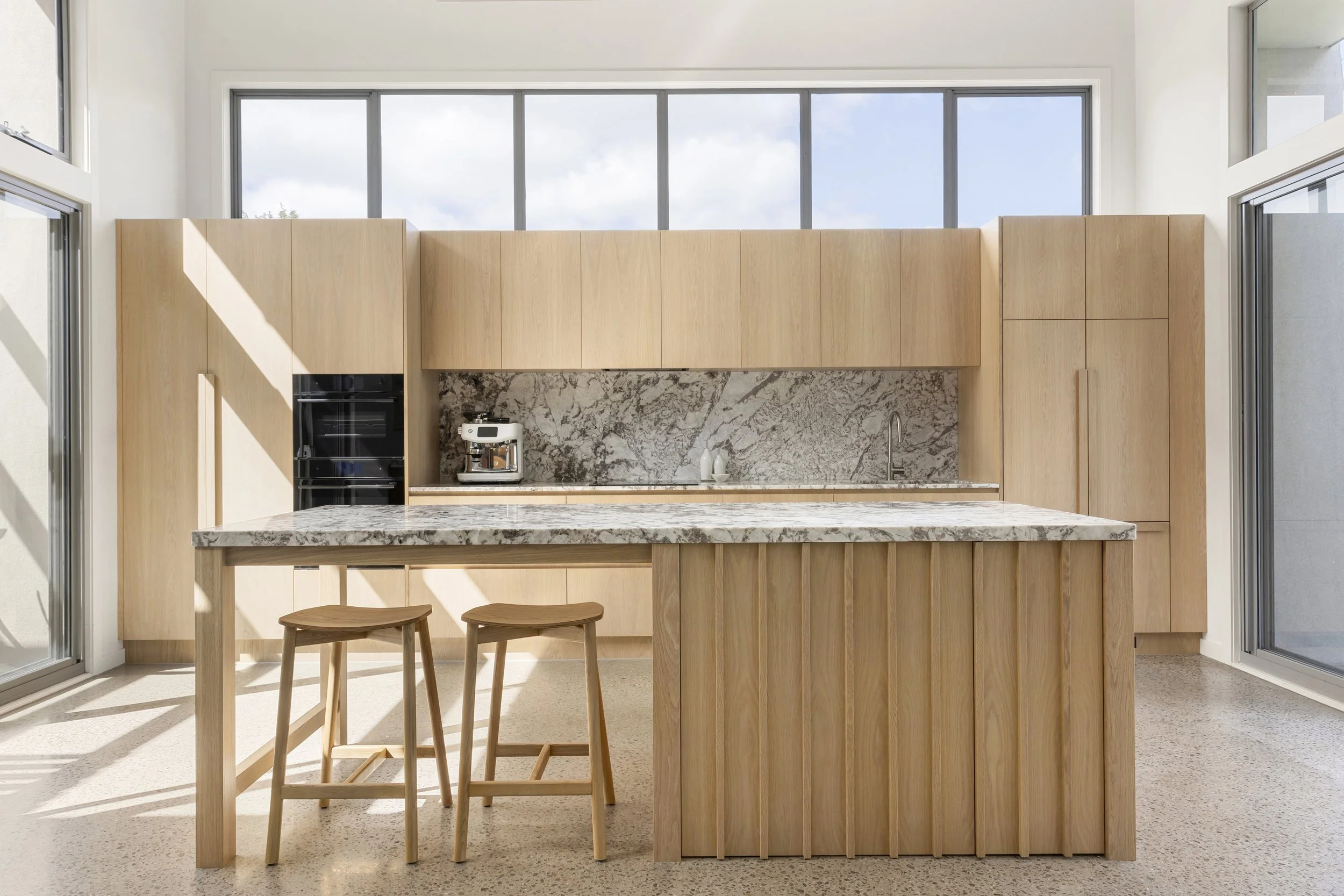 Modern kitchen with wooden cabinets, marble backsplash, a coffee maker on the counter, and a marble kitchen island with two wooden stools, large windows allowing natural light.