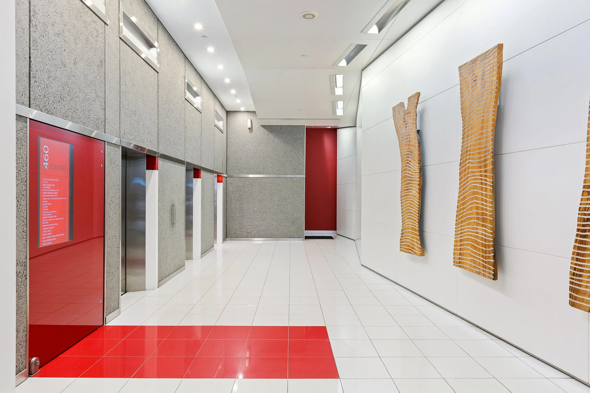 Elevator lobby with three elevators, a red informational sign, white tiled floor with red accents, a textured grey wall, and two wooden sculptures on the white wall.
