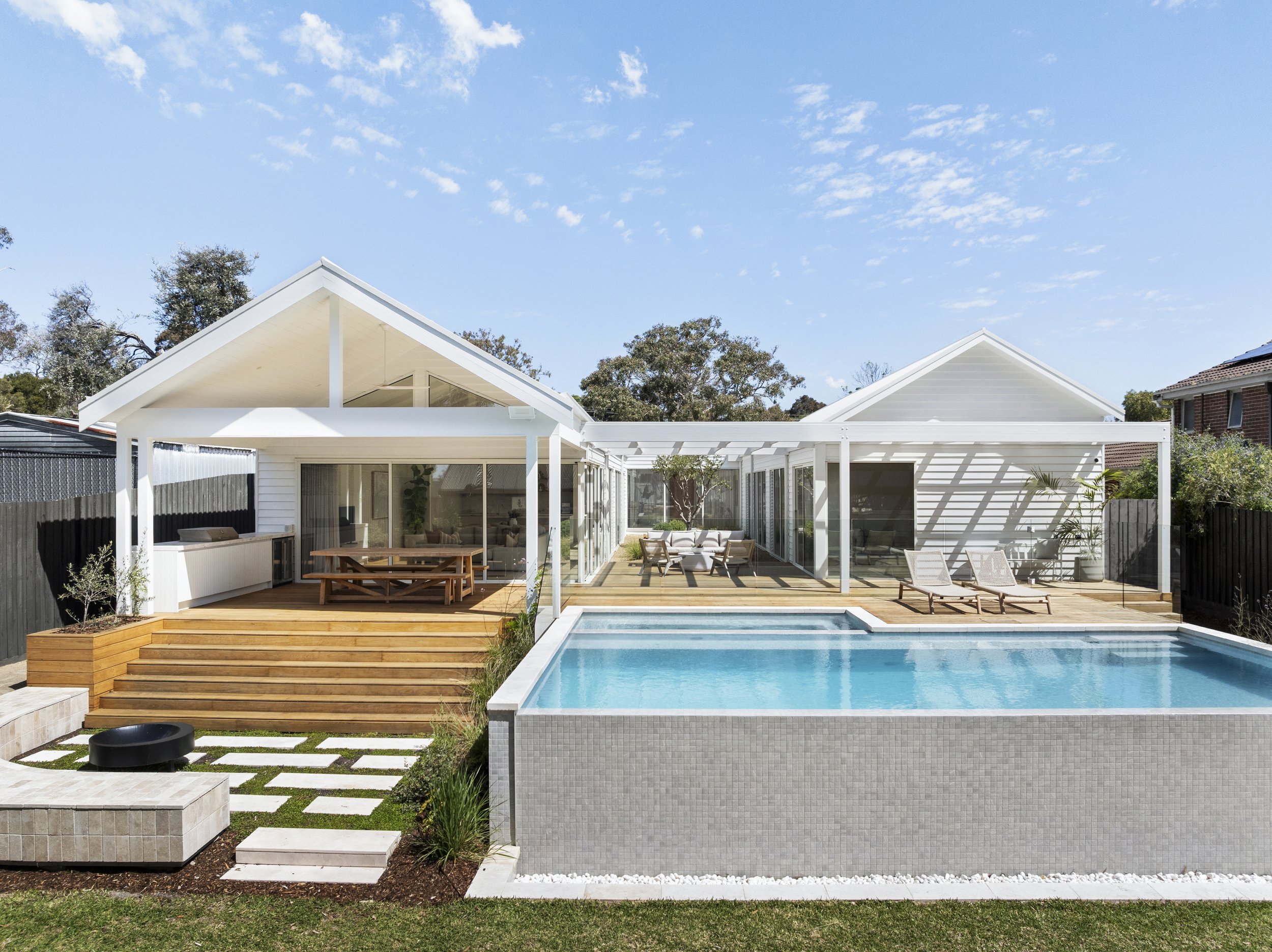 Modern backyard with a swimming pool, wooden decking, outdoor seating area, and a white house with a pergola, surrounded by trees and clear blue sky.
