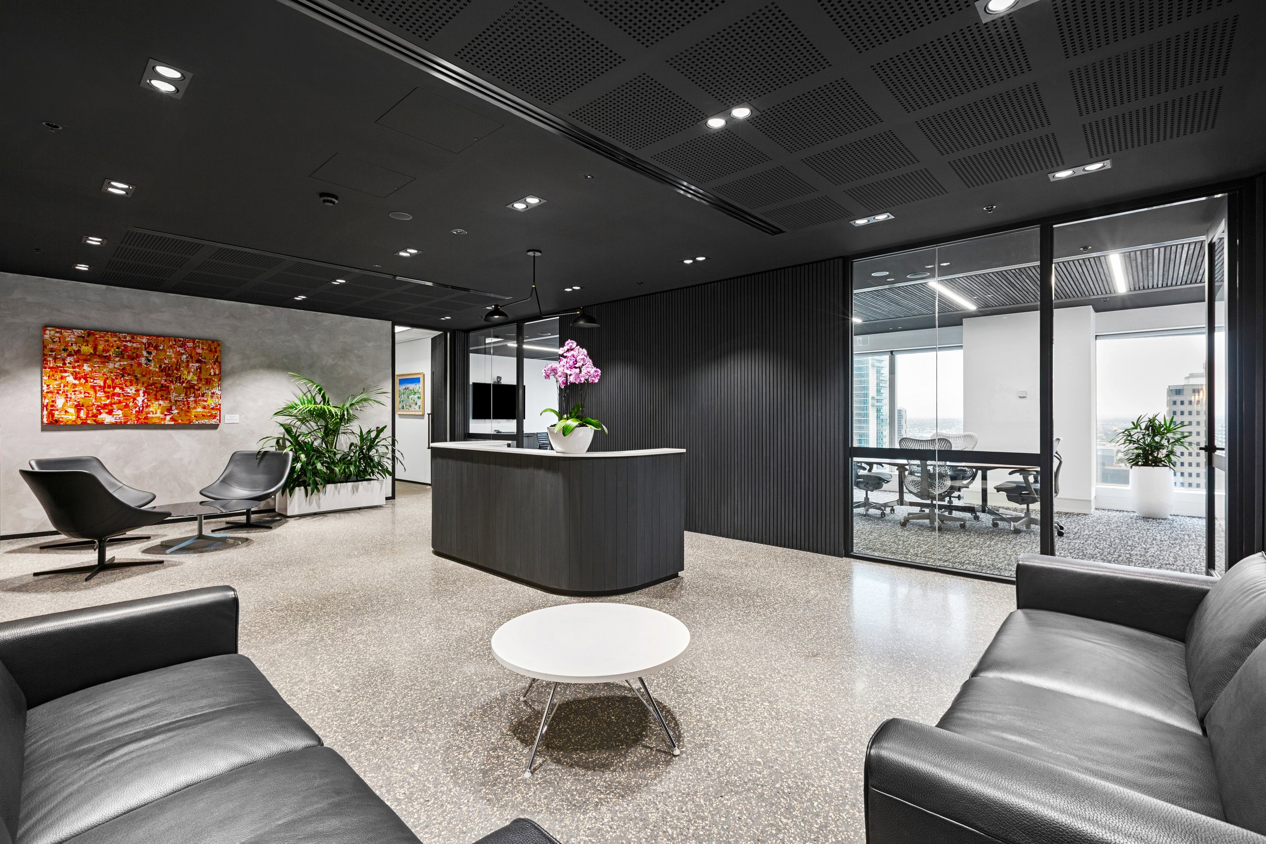 Modern office waiting area with black leather sofas, small round white tables, green plants, and a colorful abstract painting on a concrete wall. There is a reception desk with a purple orchid, and a glass conference room with chairs and large windows showing city buildings.