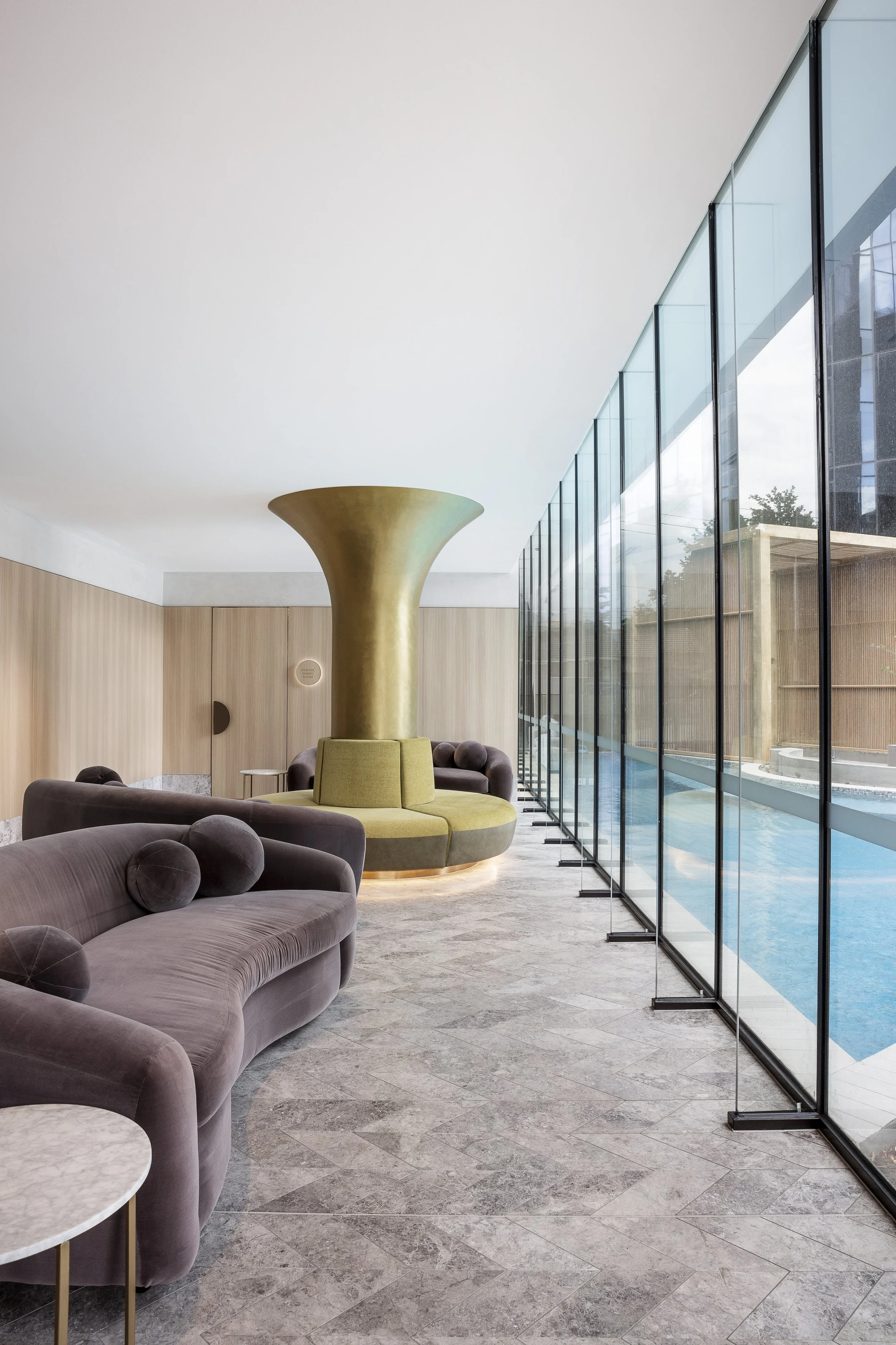 Modern lobby with purple velvet sofas, small round marble-top tables, a large gold sculptural element, and floor-to-ceiling windows overlooking a pool area.