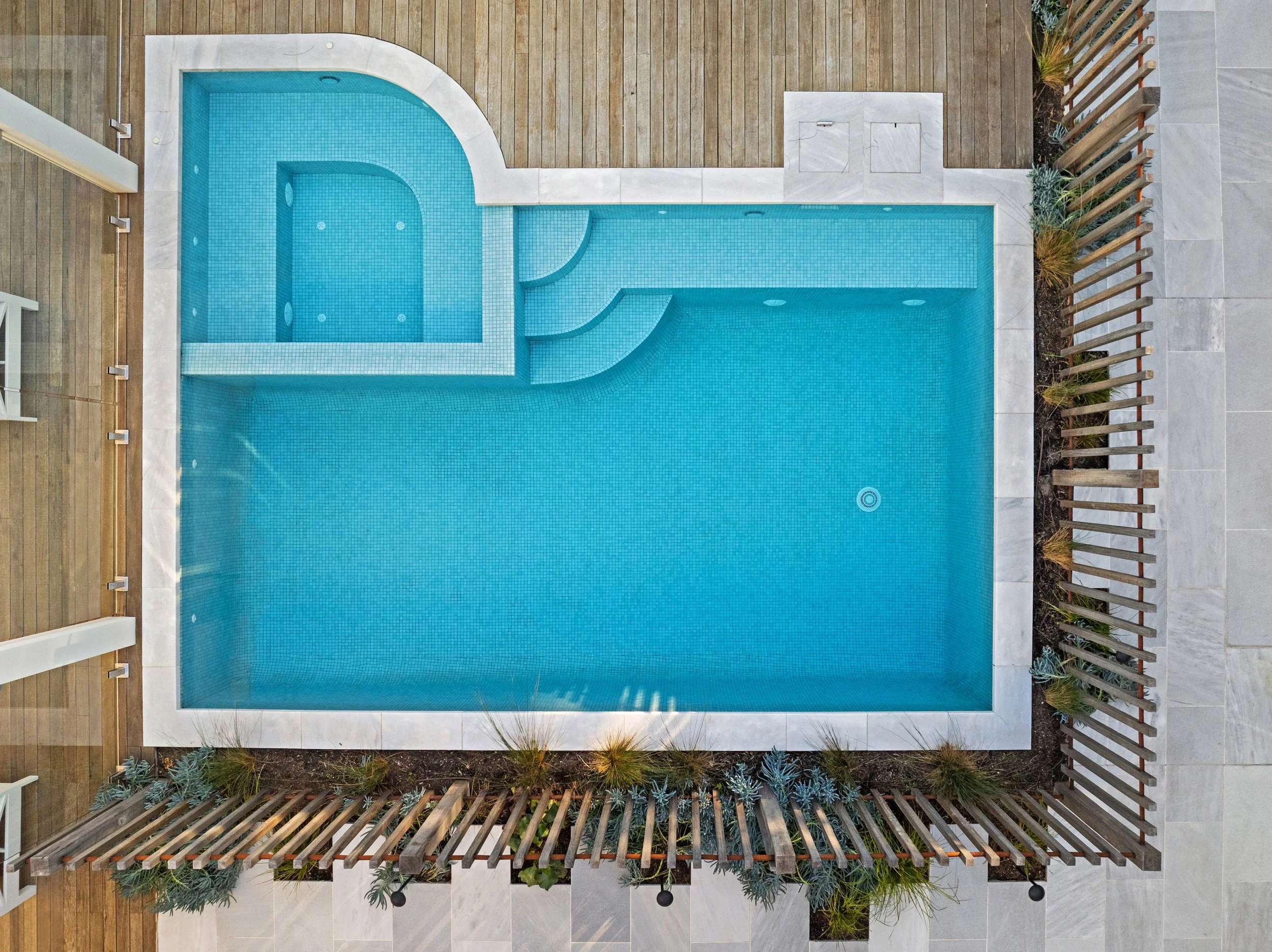 Top-down view of a rectangular swimming pool with a smaller curved hot tub attached, surrounded by a wooden deck and a gray tiled patio area, enclosed by a fence with plants.