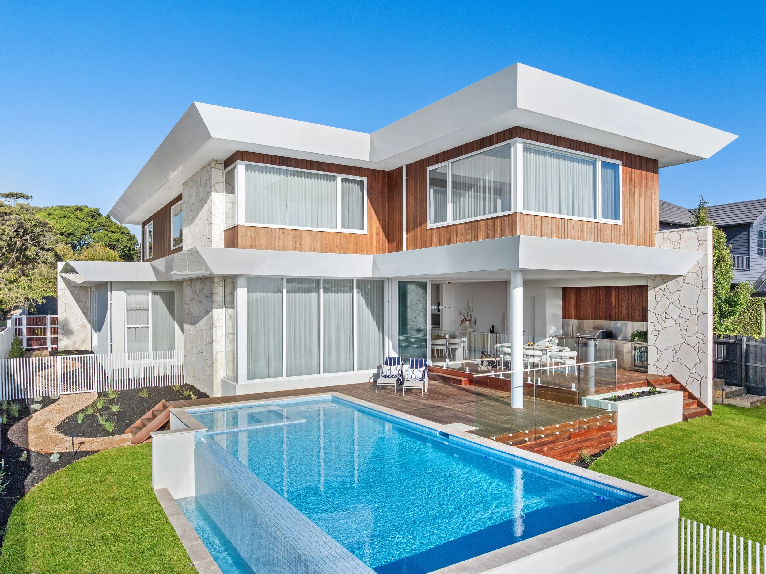 Modern two-story house with large windows, wooden and stone exterior, and a flat white roof. In the foreground, there is a swimming pool with a clear blue surface, surrounded by a wooden deck and green lawn. The backyard features outdoor patio furniture and a barbecue grill, under a sunny sky.