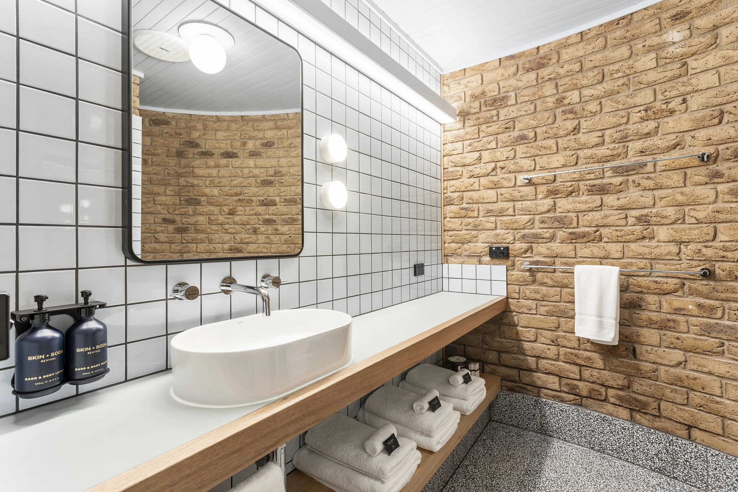 Modern bathroom with a large rectangular mirror, white subway tile walls, wooden vanity with a white sink, black soap and lotion bottles, and towels neatly folded on open shelves. Exposed brick wall adds rustic charm. There's a towel bar with a white towel and a terrazzo floor.