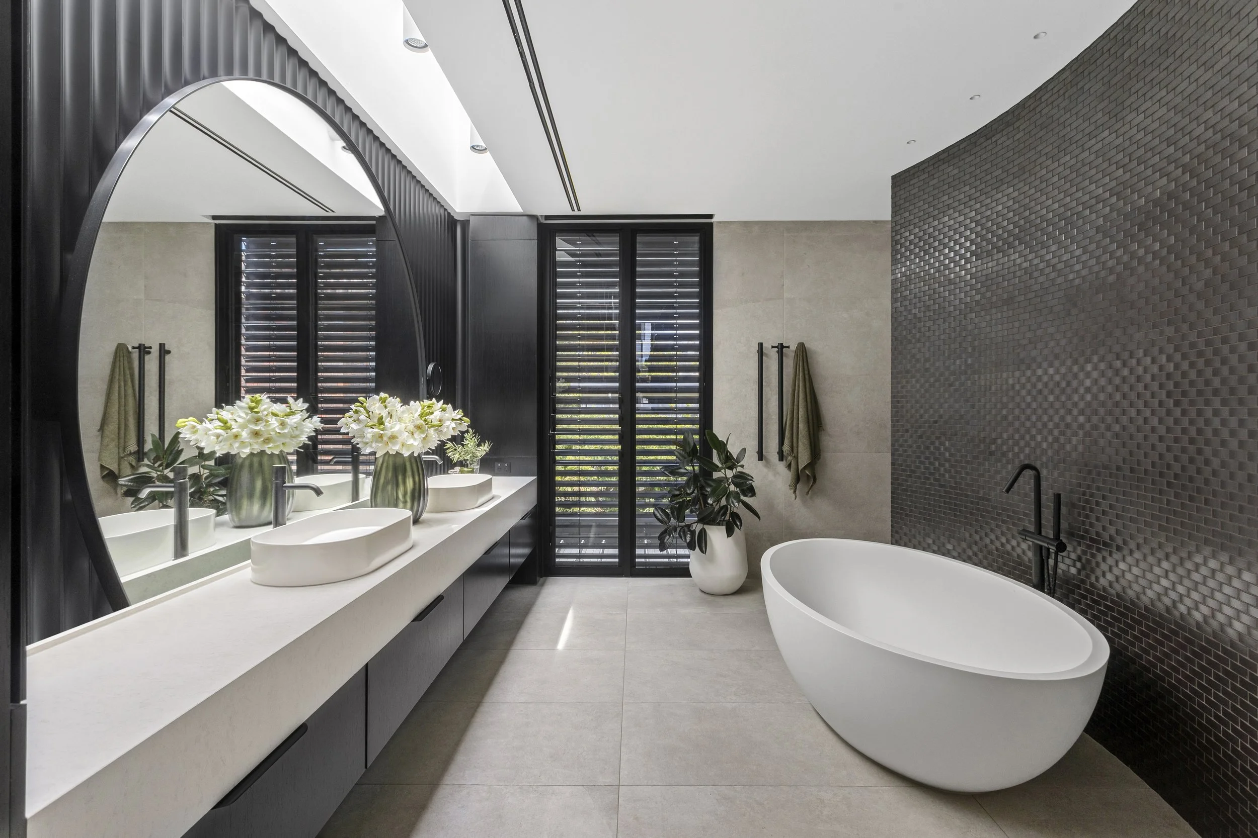 Modern bathroom with a freestanding bathtub, black textured wall, double sinks with flower vases, black window shutters, and minimalist decor.