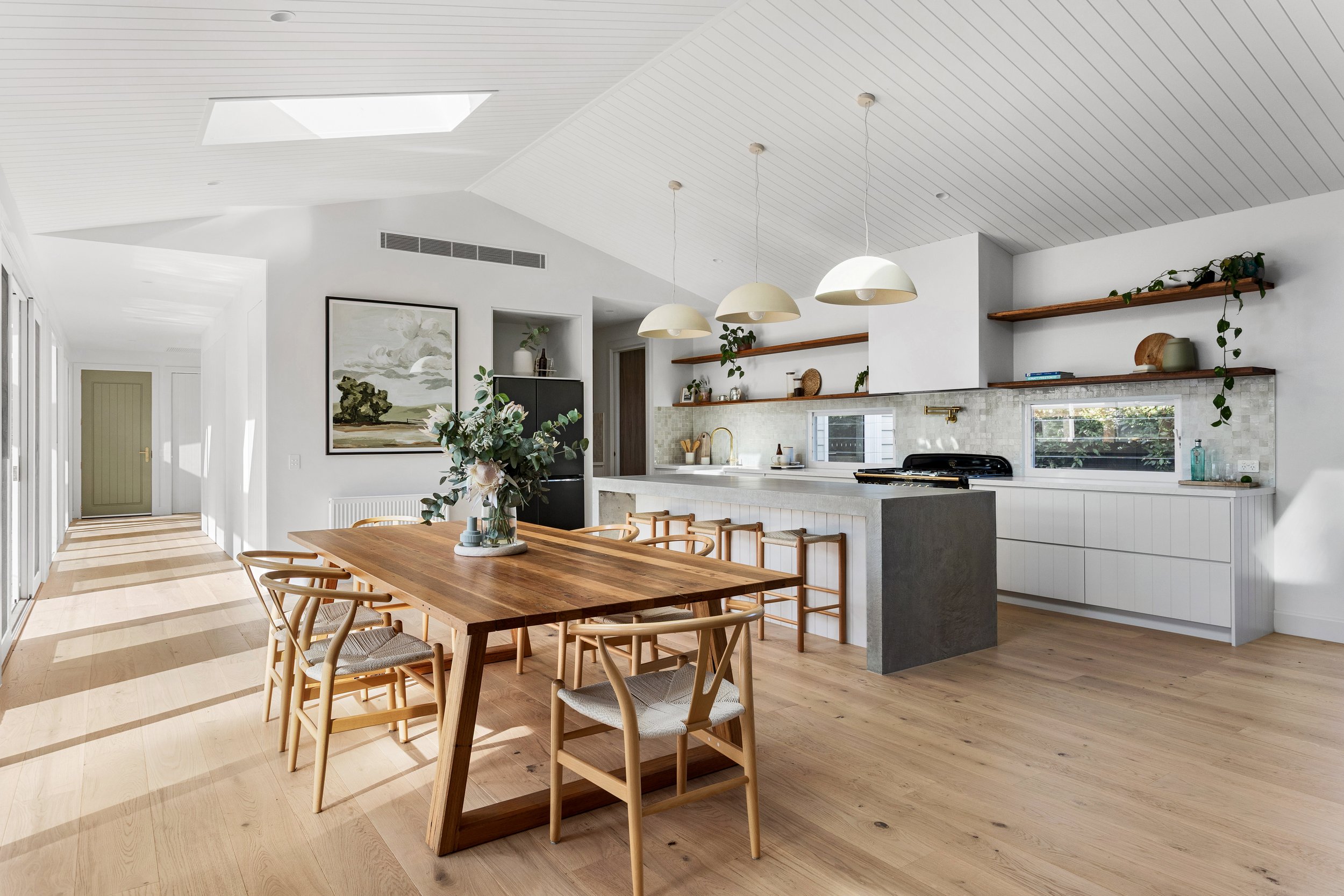 A modern, open-concept kitchen and dining area with white walls and ceiling. There is a large wooden dining table with chairs, a kitchen island with stools, suspended pendant lights, and open shelves with decor items. Natural light streams through a window, and the space features minimalistic decor with plants.
