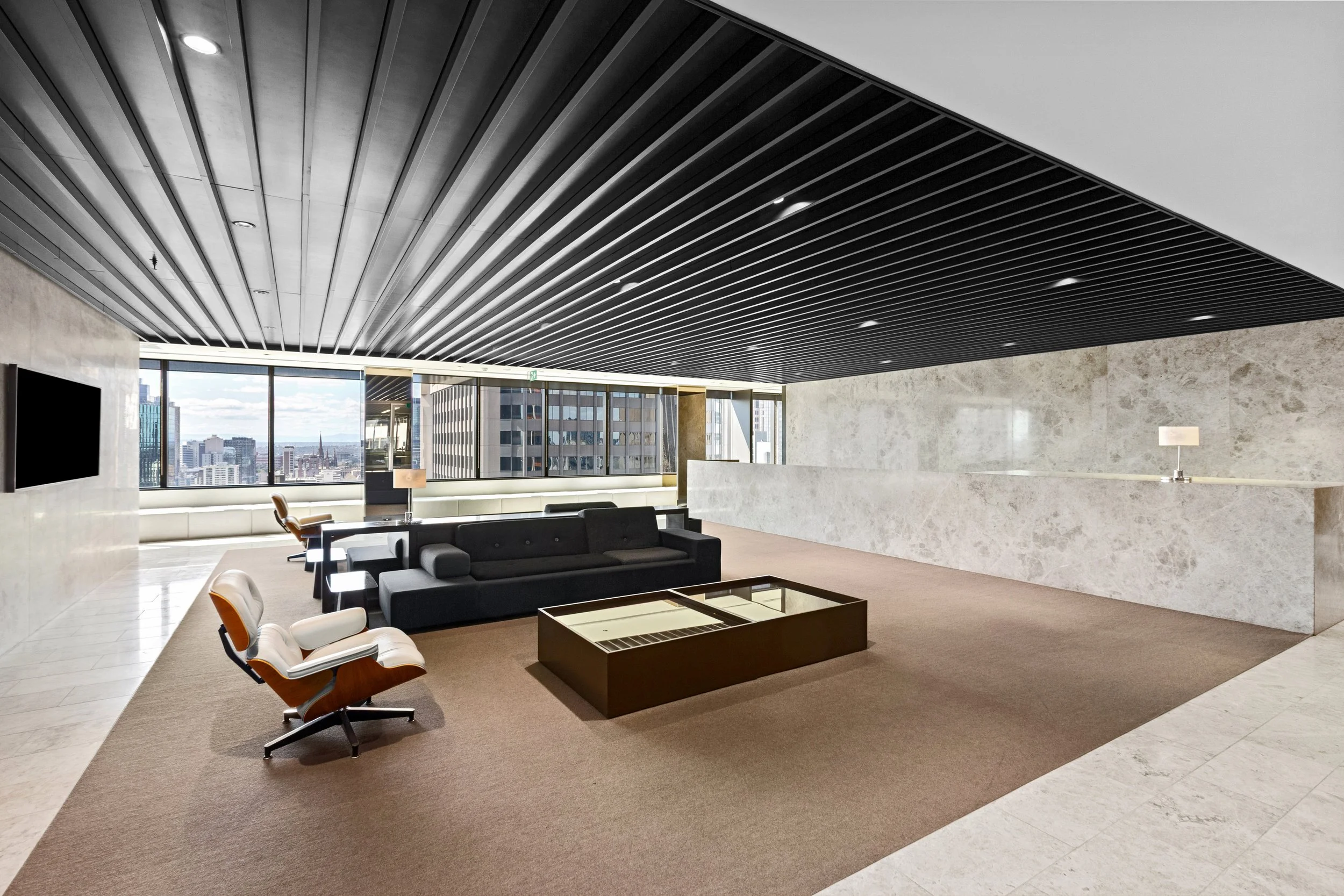 Modern office lounge area with black sofa, white chairs, coffee table, large windows, city view, and a black ceiling with linear lights.