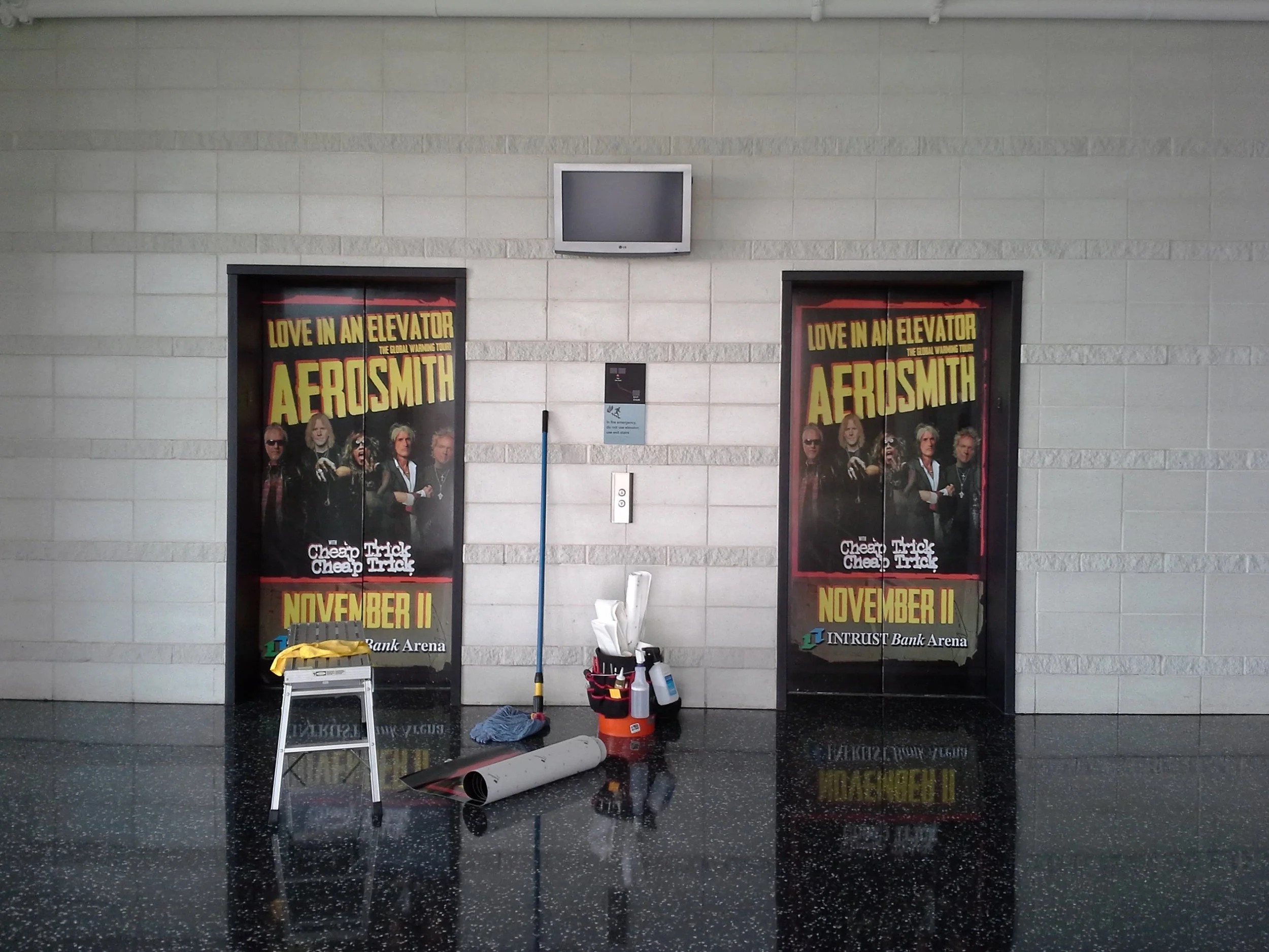 Elevator doors with posters for Aerosmith's tour and various cleaning tools and supplies in front, on a shiny black floor at an indoor location.