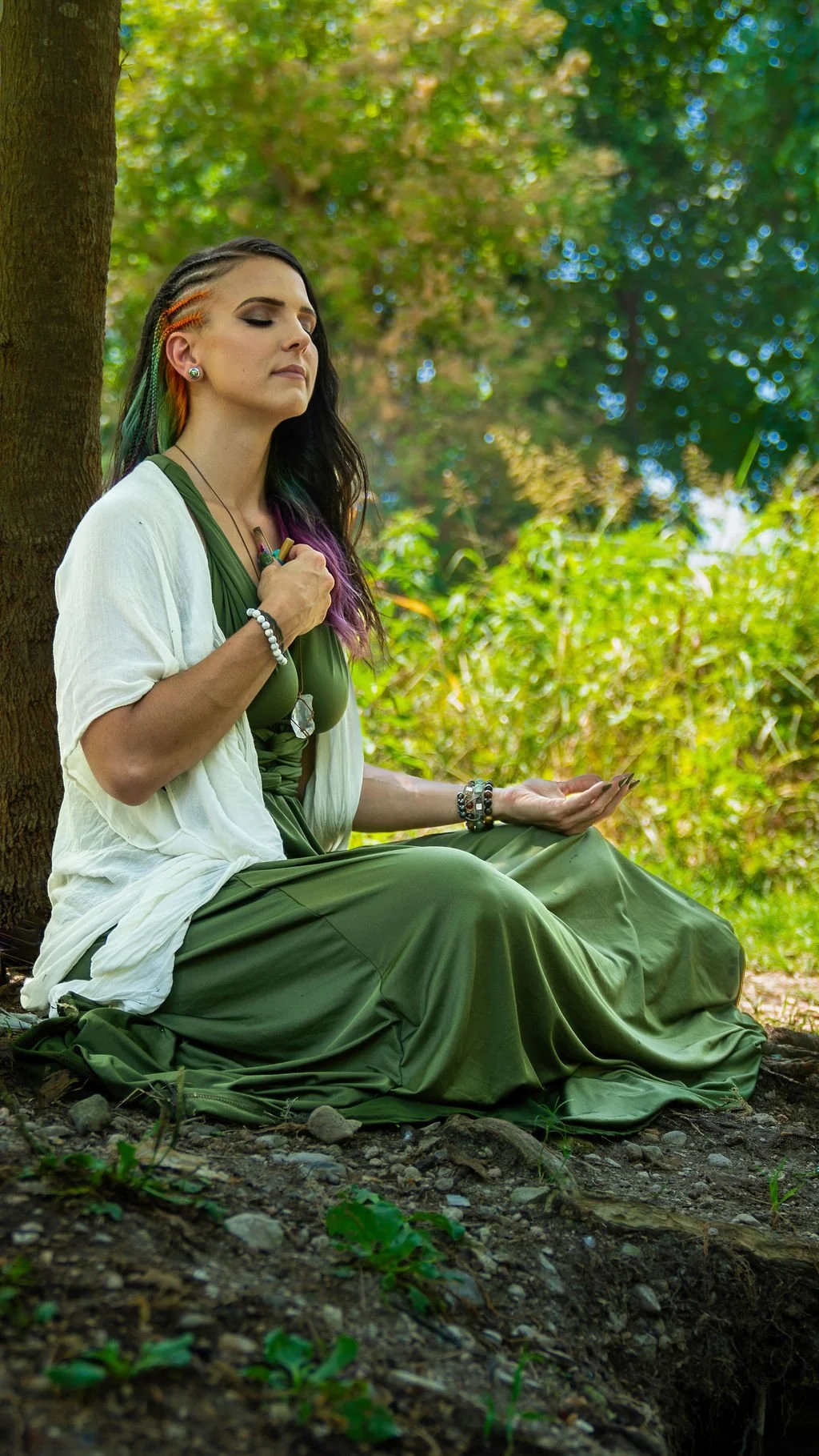 A woman practicing meditation outdoors, sitting cross-legged under a tree with her eyes closed and hands resting on her lap.