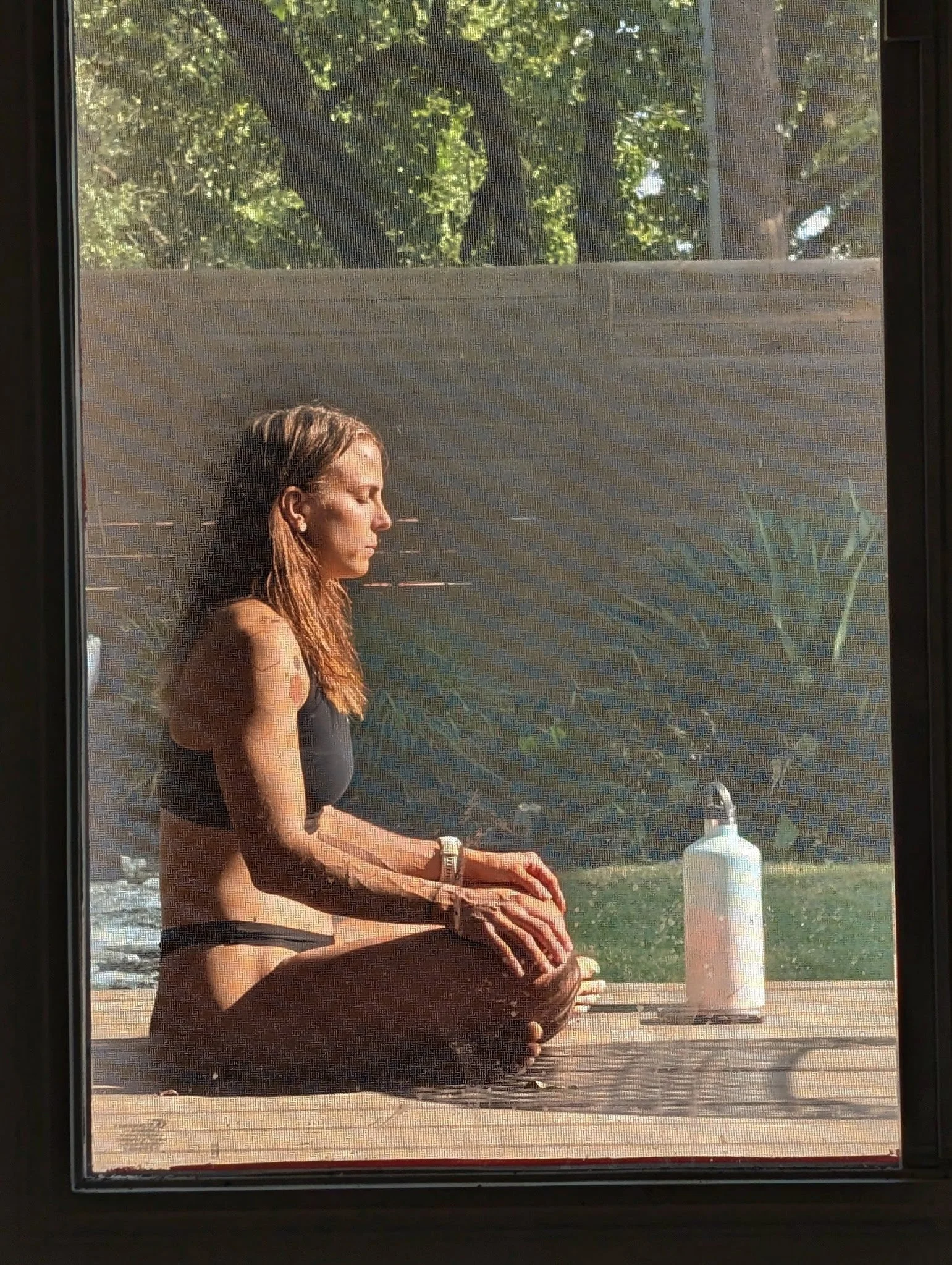 A woman sitting cross-legged on a wooden deck outside, practicing yoga or meditation, with a spray bottle or sanitizer nearby, seen through a window screen.
