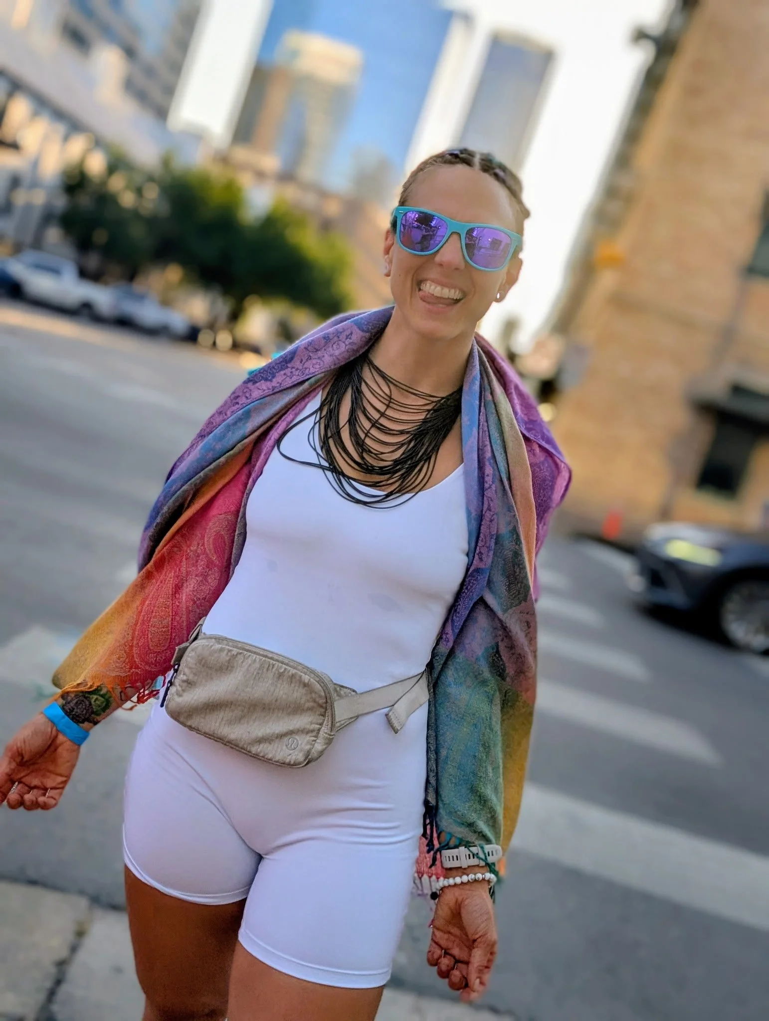 Smiling woman with sunglasses, a rainbow-colored shawl, white athletic wear, and a beige waist bag, standing on city street with buildings in background.