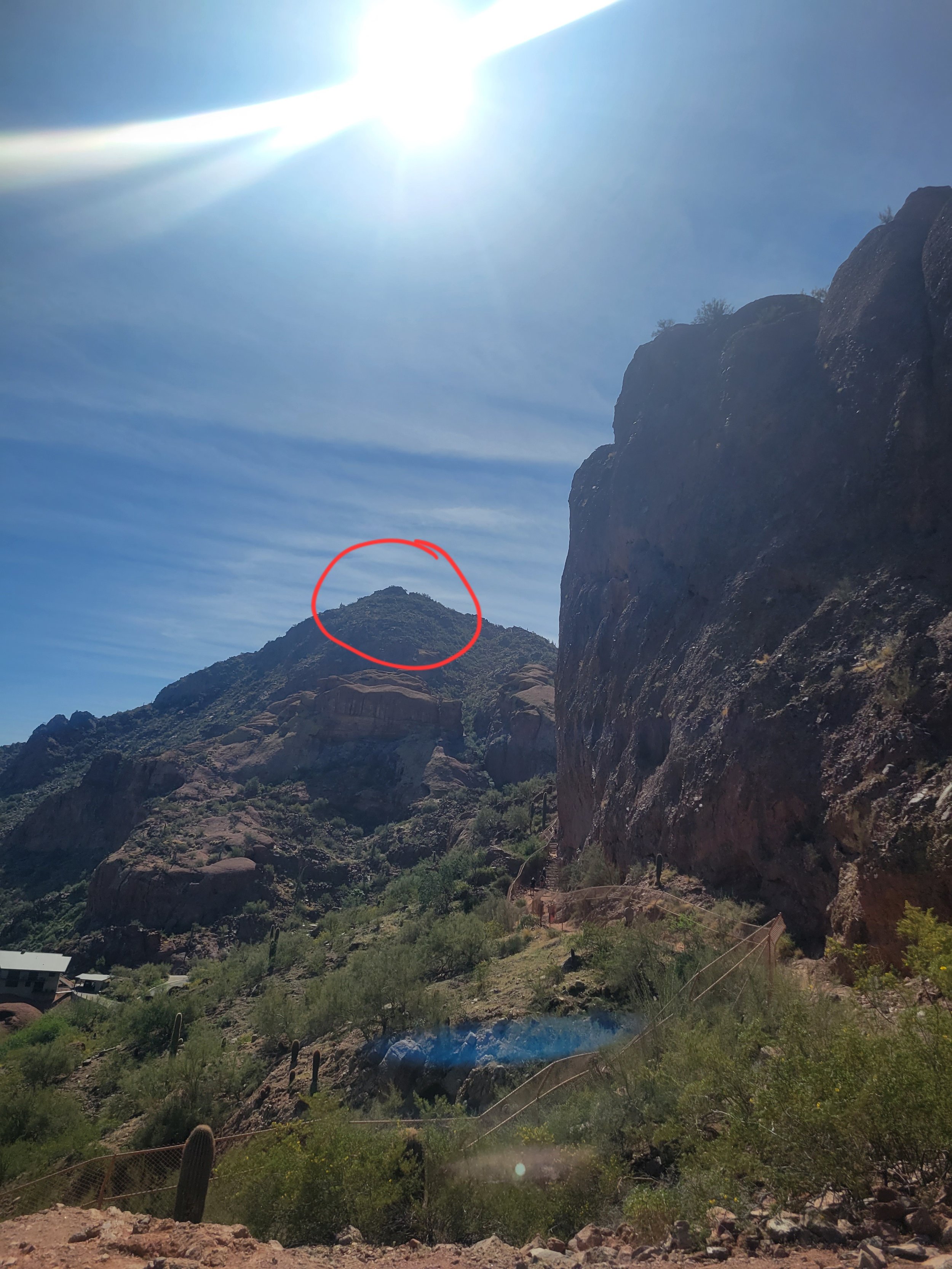 A mountainous landscape on a sunny day with bright sunlight, a blue sky with wispy clouds, and green desert vegetation. A red circle highlights a peak in the mountain.